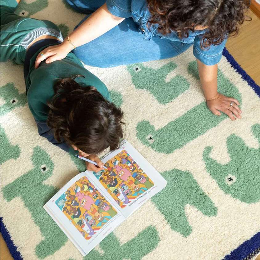 Bambino che sfoglia un libro illustrato sdraiato su un tappeto avorio con motivi verdi