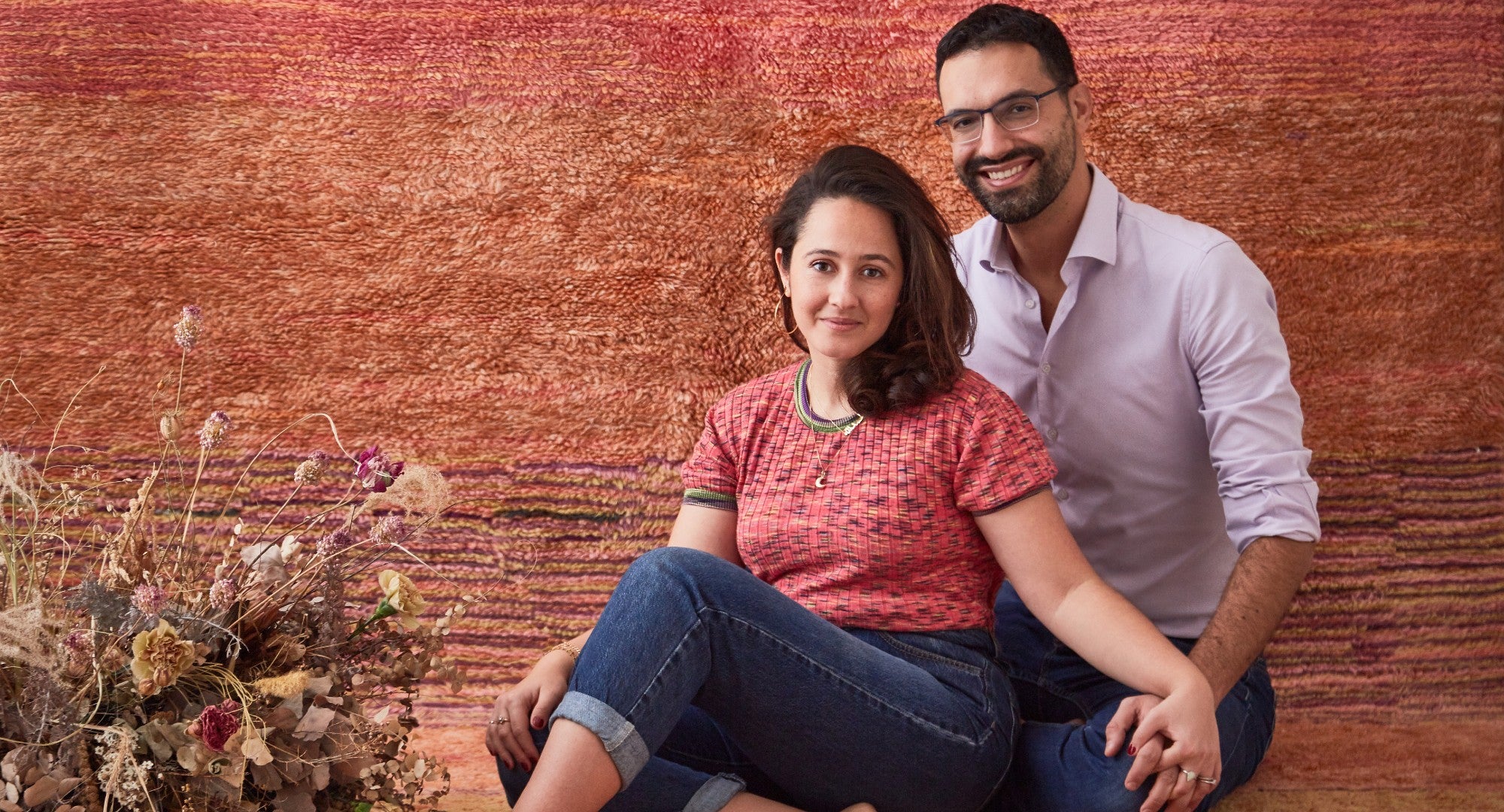 coppia sorridente seduti su un tappeto dai toni rossi e rosa che fa anche da sfondo. ai loro piedi è presente una decorazione floreale