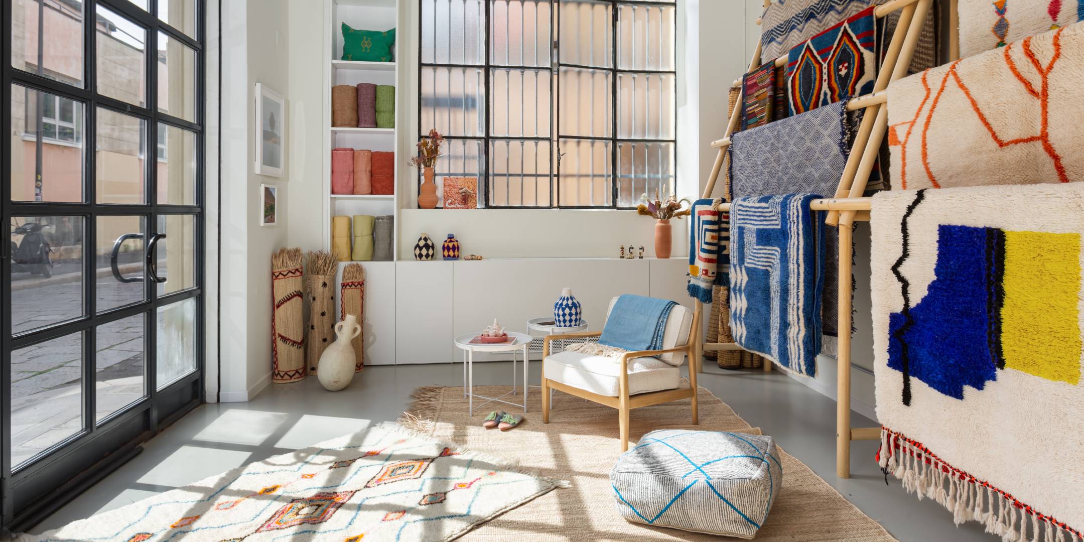 Interno di Casa Amar, uno showroom di tappeti marocchini a Milano. Spazio luminoso e arioso che espone colorati tappeti e tessuti marocchini. Grandi finestre illuminano un'elegante area espositiva con una poltrona in legno, cuscini da pavimento decorati e un espositore per tappeti. Scaffali con rocchetti di filo e oggetti decorativi evidenziano l'autentico artigianato marocchino in questa curata boutique milanese.