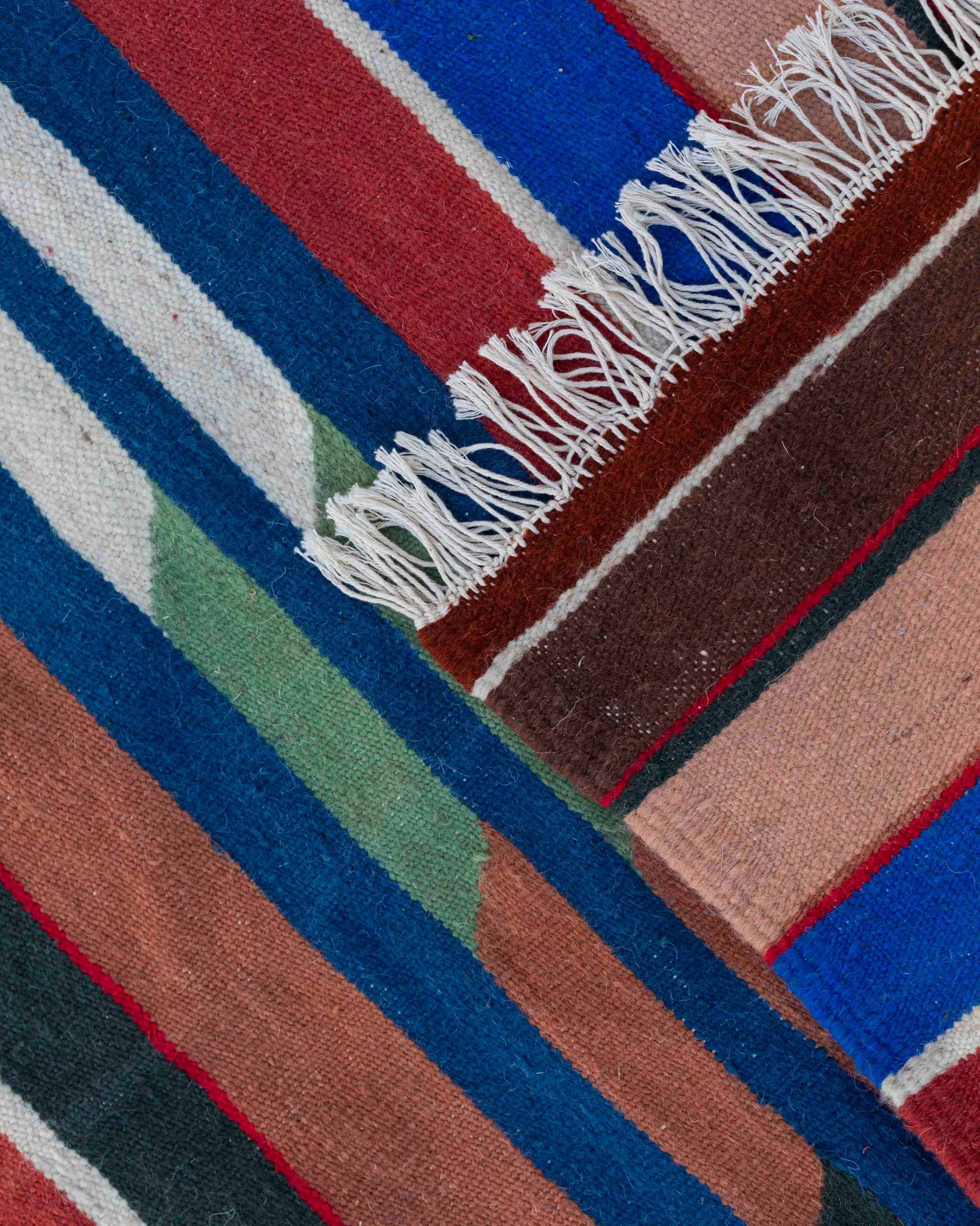 Dettaglio delle frange sottili e delle righe blu, rosse, marroni e rosa del kilim Hanbel in lana