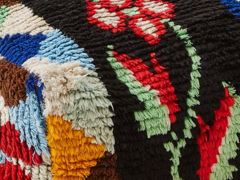 dettaglio di un pouf ricavato da un vecchio tappeto. su lato sinistro sono presenti una seria di triangoli colorati e al centro un fiore con petali rossi e bianchi su sondo nero