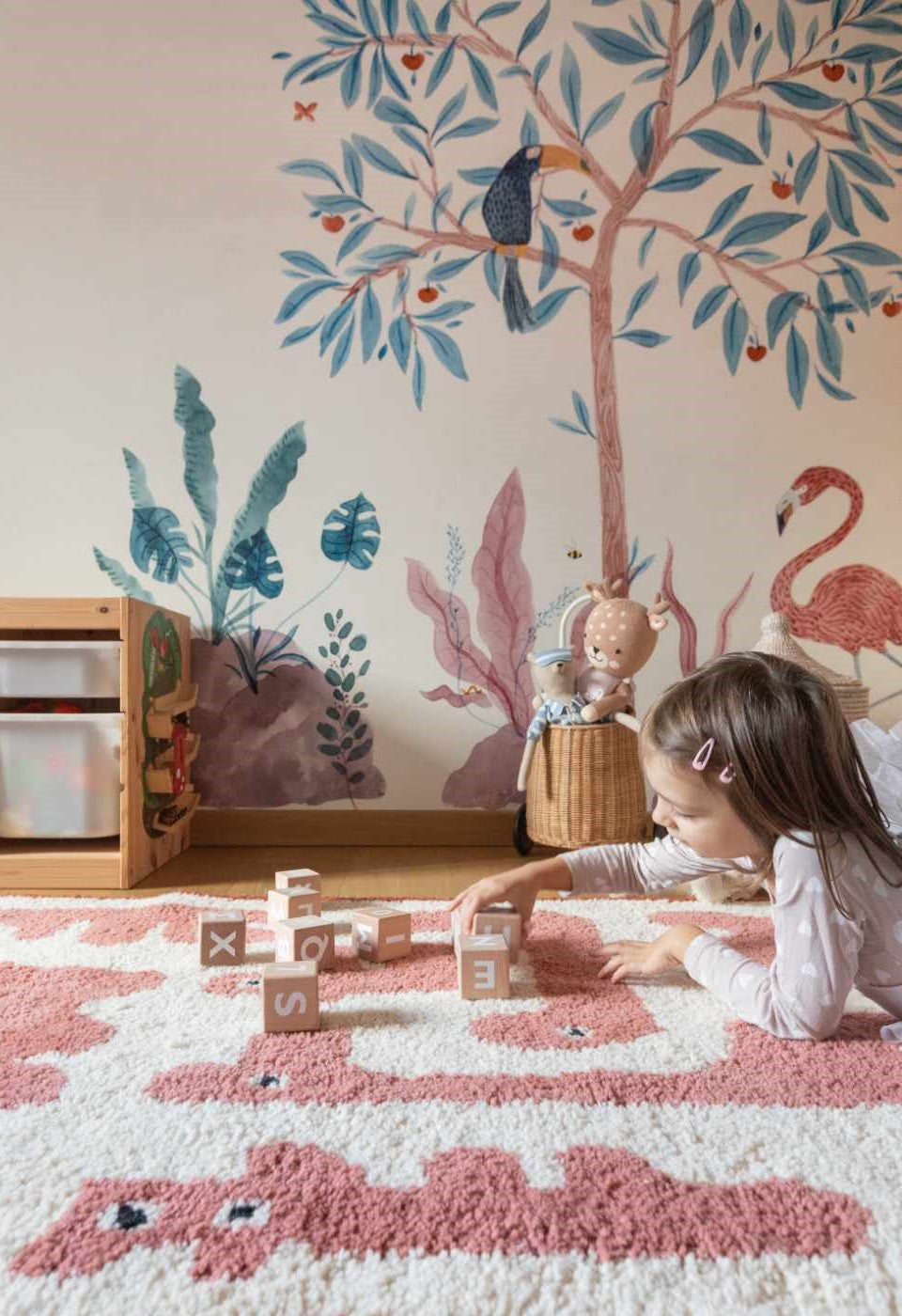 Cameretta con tappeto rosa e bianco decorato con motivi di animali. Una bambina gioca con dei cubi sul tappeto morbido, sullo sfondo un murale tropicale con fenicottero e tucano, e un mobile contenitore in legno.