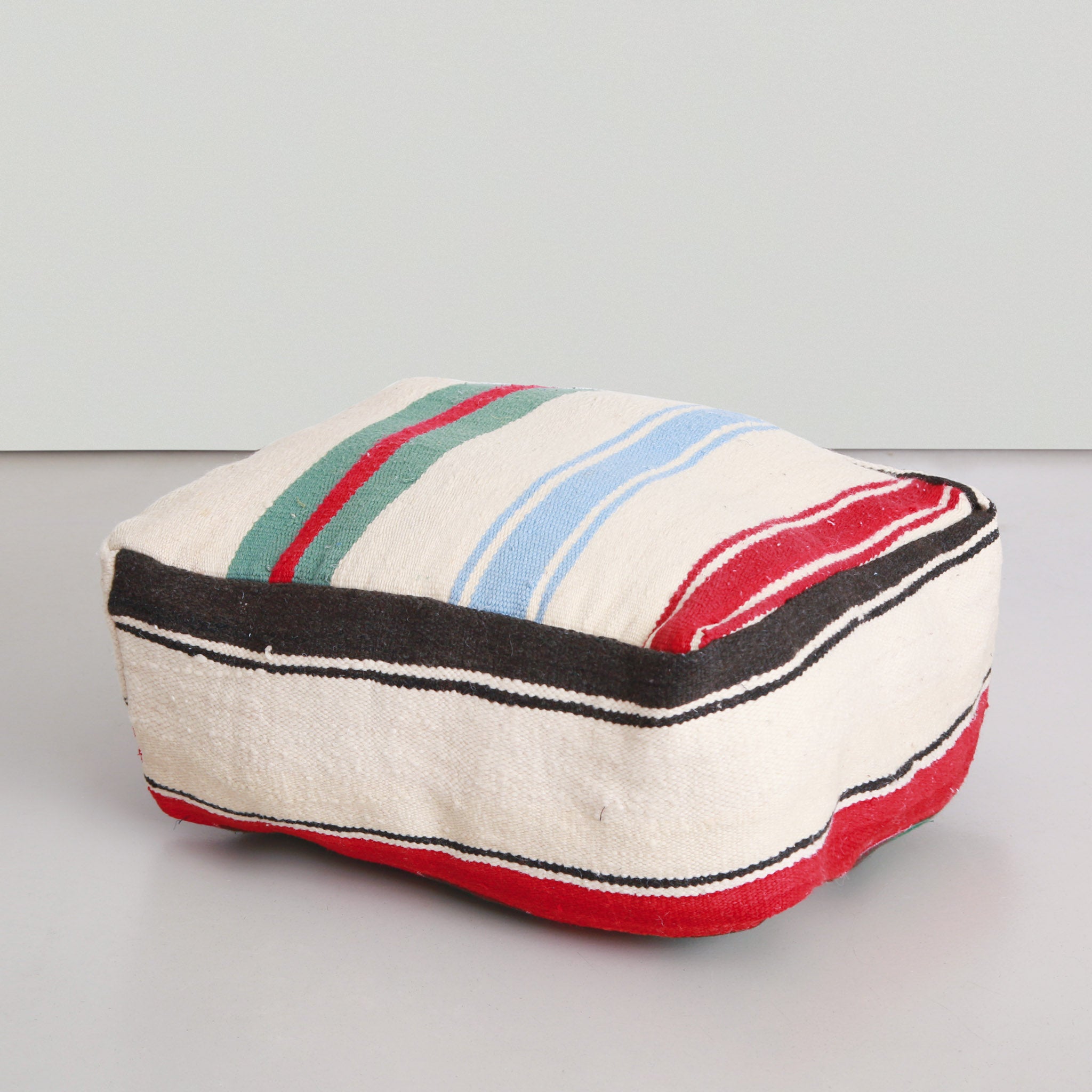 Pouf marocchino quadrato realizzato con coperta vintage in lana, con motivo a strisce colorate bianche, rosse, verdi e azzurre su sfondo chiaro