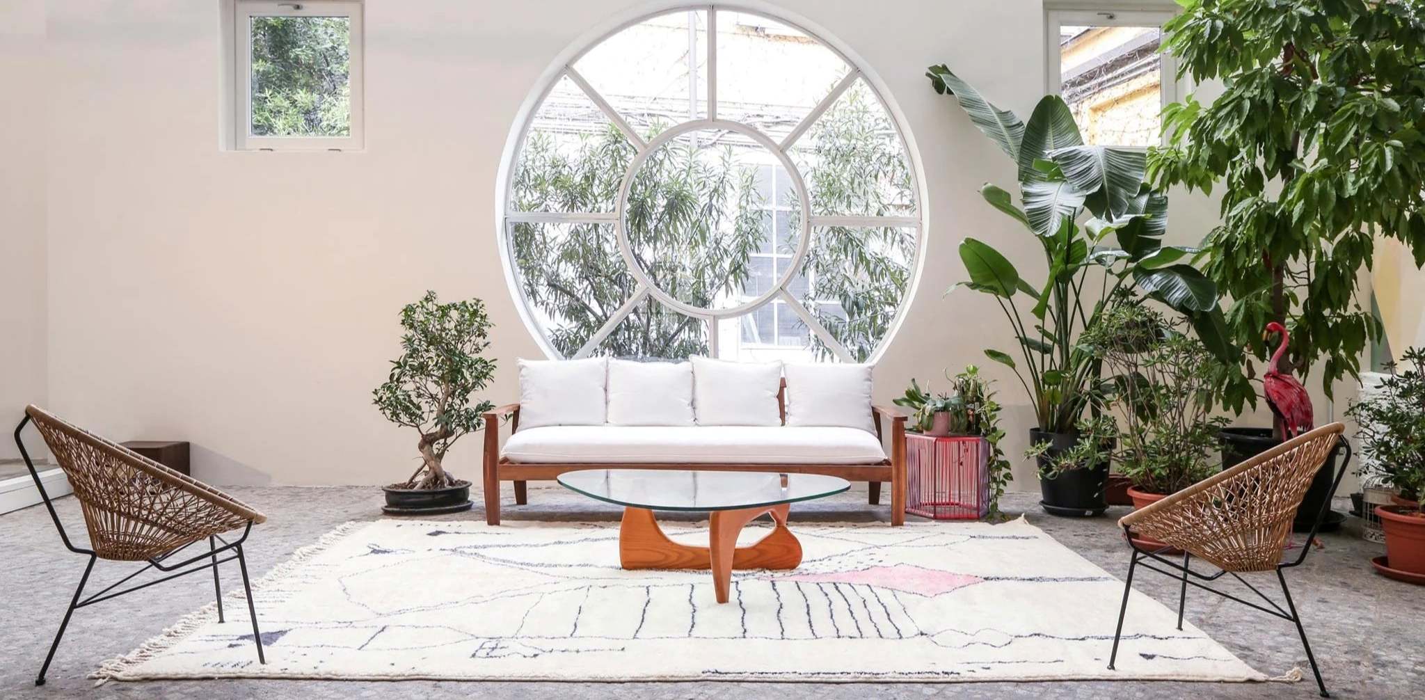 Tapis berbere Mrirt authentique en laine naturelle sublimant un studio d'architecture contemporain. Design minimaliste avec canapé blanc, fauteuils en rotin et table Noguchi, magnifié par une fenêtre circulaire et végétation méditerranéenne