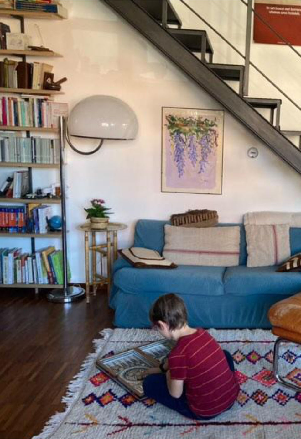 Salon confortable avec un tapis vintage marocain aux motifs géométriques colorés, où un enfant joue à un jeu de société. La pièce comprend un canapé bleu, des étagères et un escalier métallique.