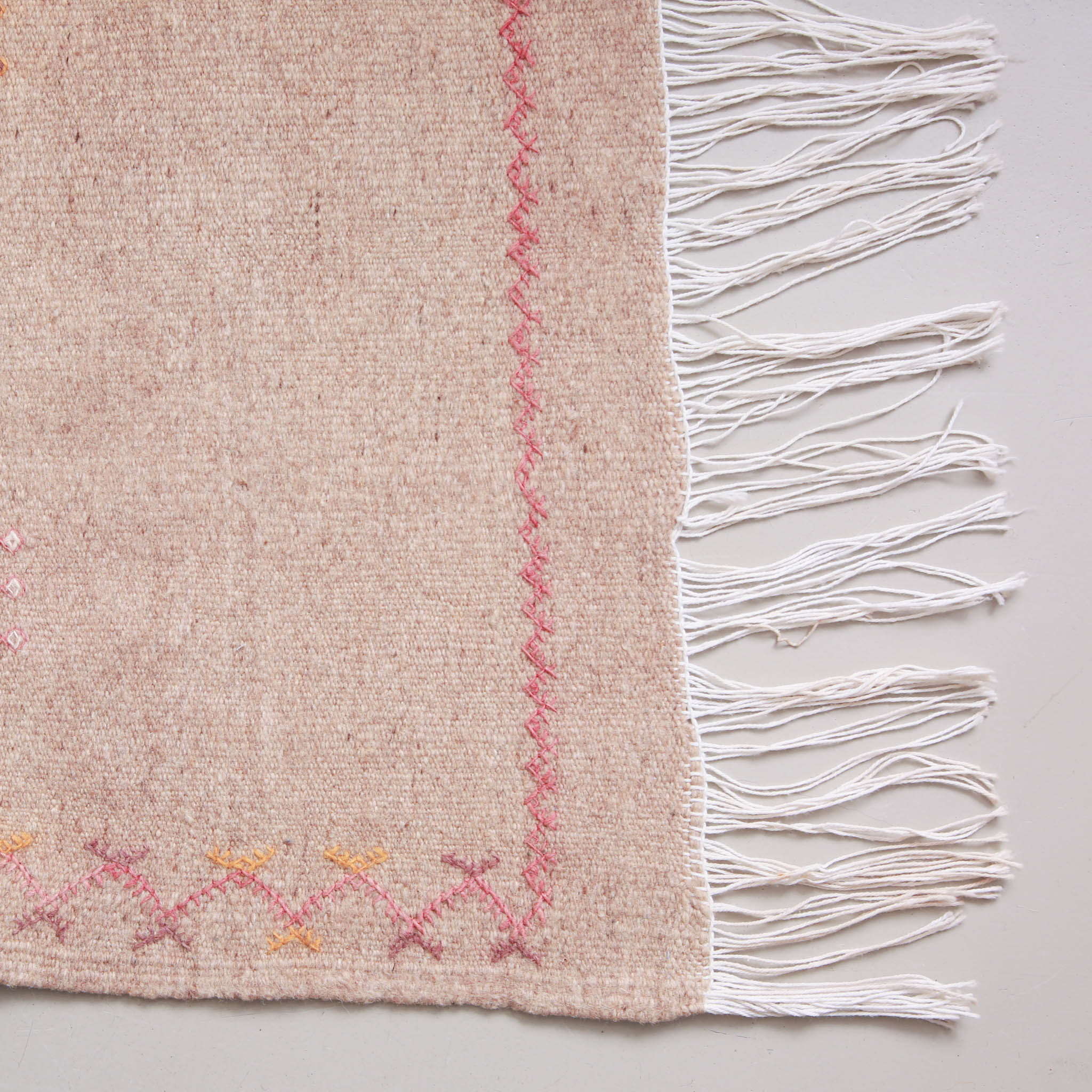 Dettaglio angolo tappeto marocchino in tinta rosa cipria con ricami geometrici e frange intrecciate, bordatura decorativa artigianale