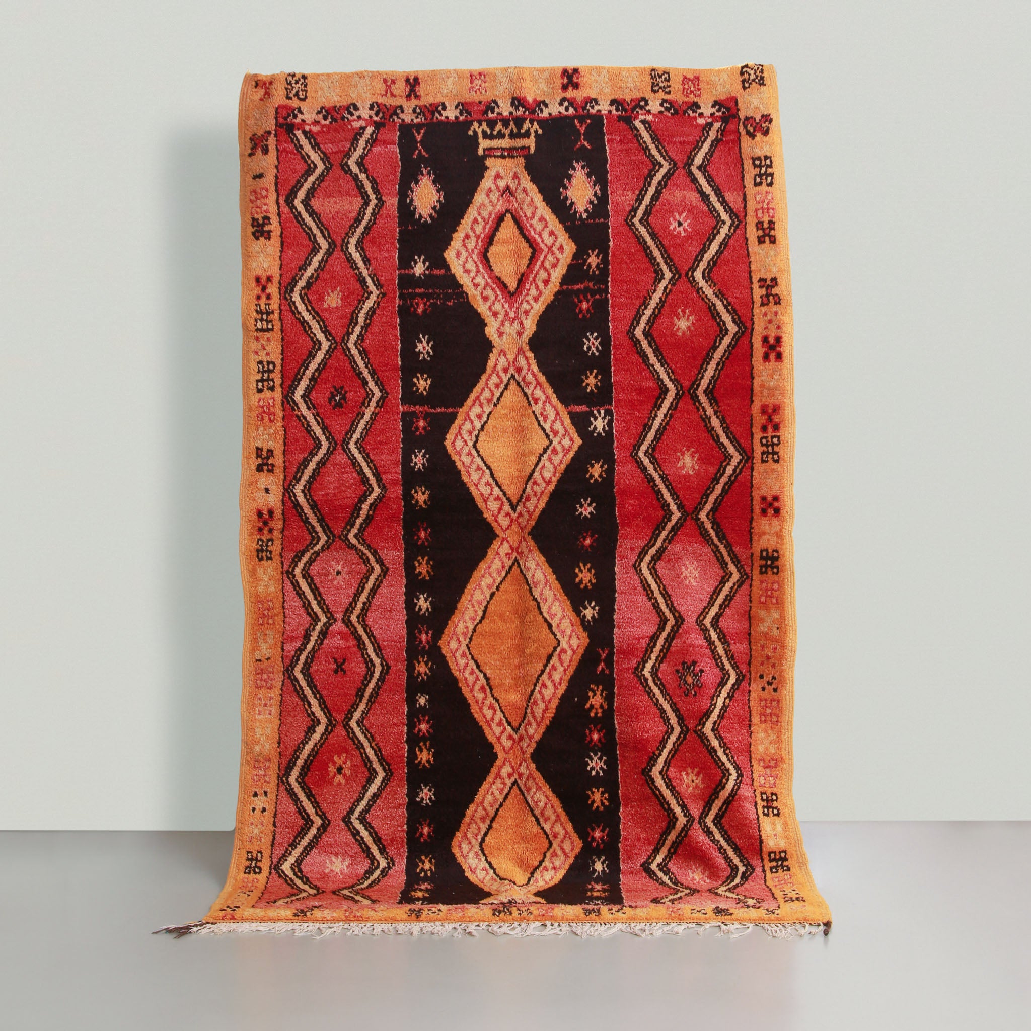 tappeto in lana molti colore rossa nera arancione giallo. nella colonna centrale nera ci sono 4 rombi gialli arancioni con simboli, le due colonne laterali sono rosse con linees spezzate nere e bianche. la cornice è gialla e arancione con tanti simboli