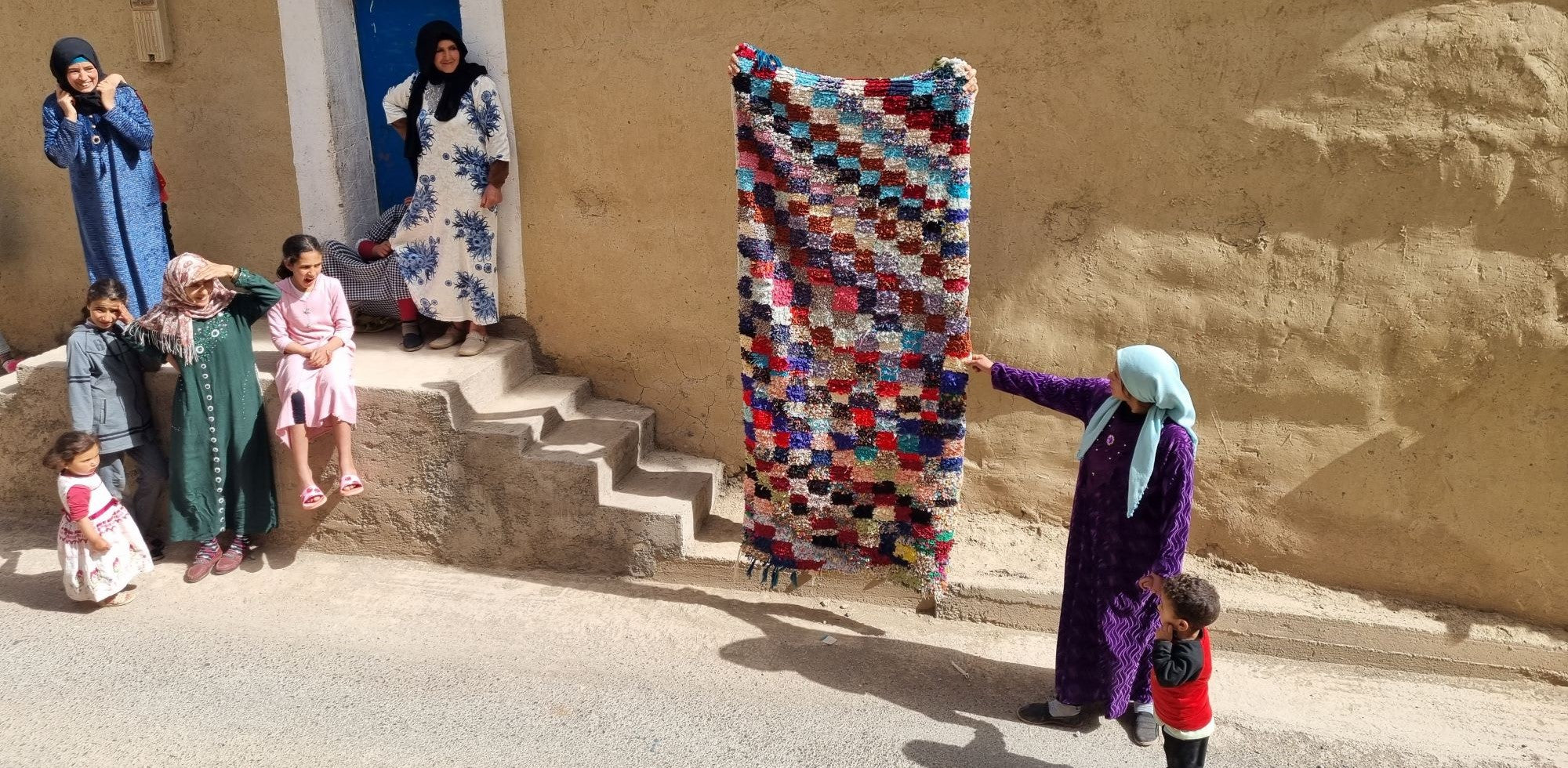 tappeto boucherouite appeso sul marciapiede di una strada di montagna mentre le donne del villaggio lo osservano
