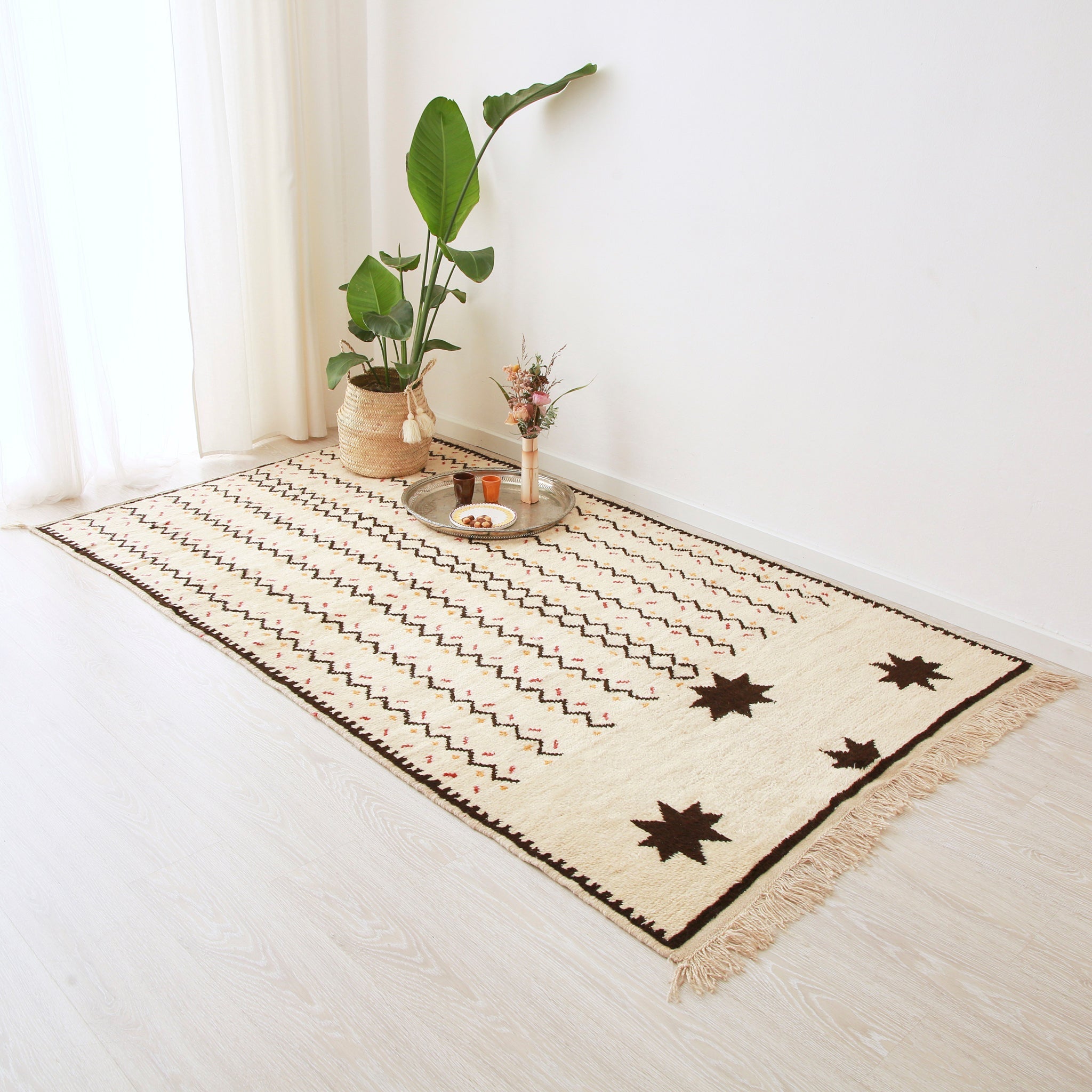 tappeto stile azilal disteso realizzato su base bianche con linee a zig zag nere e quattro stelle nere, racchiso in un bordo nero.sul tappeto sono presenti un pianta e un piatto in argento con degli accessori e una composizione floreale