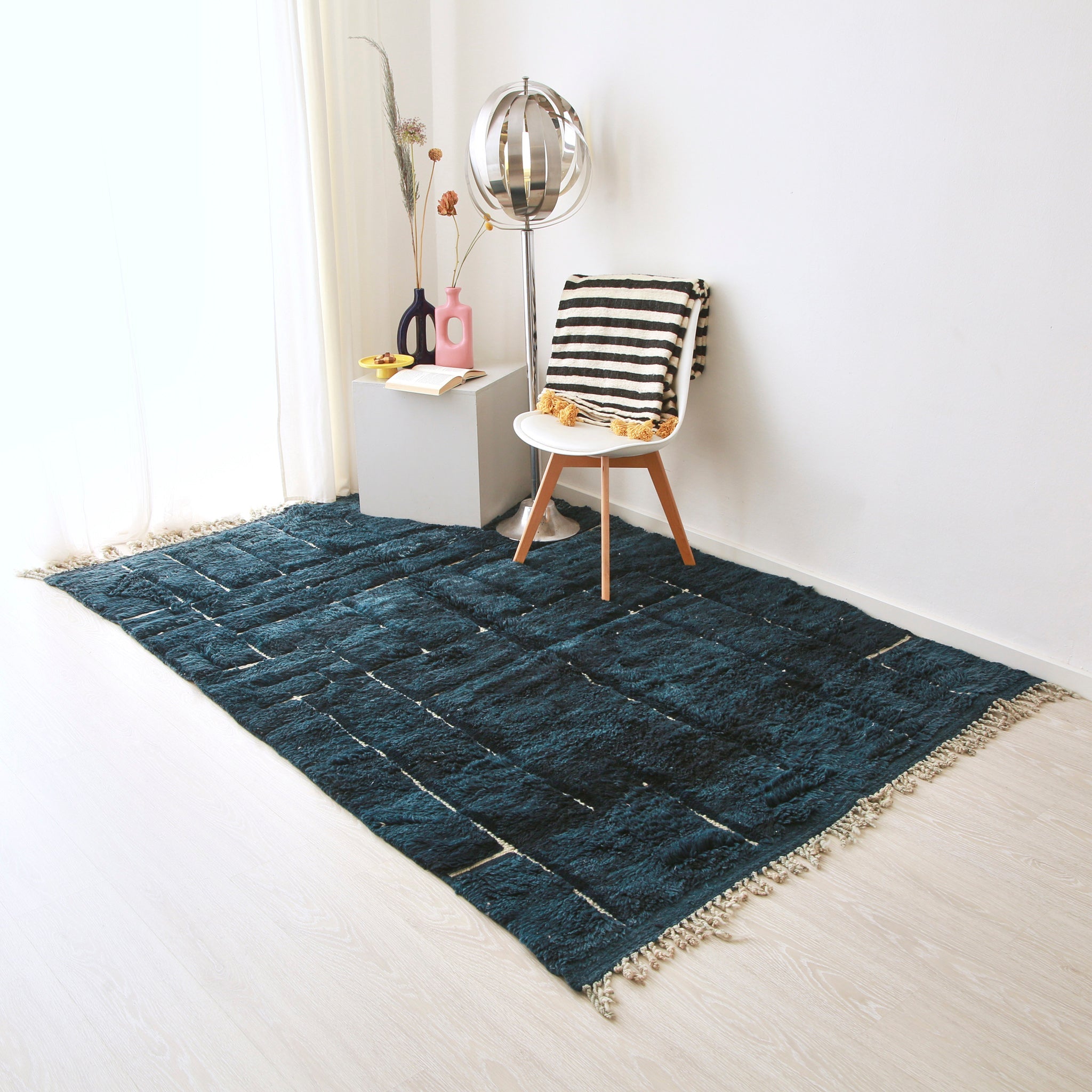 tappeto beni mrirt blu disteso sul pavimento. Sul tappeto ci sono una sedia con sopra una coperta in lana a righe bianche e nere con pon pon gialli, una lampaga in metallo stile anni 70 e delle ceramiche del marocco