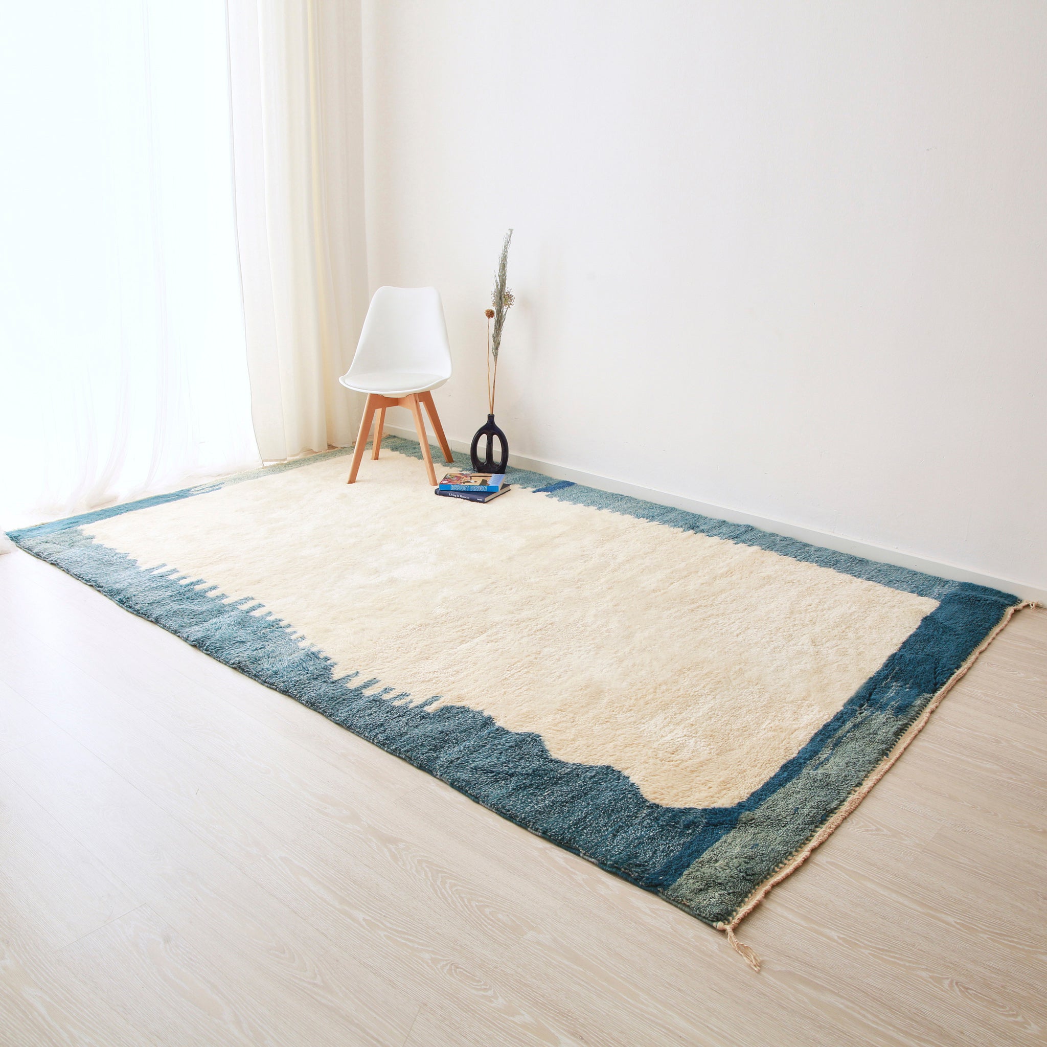grande tappeto beni mrirt realizzato in lana bianca con un bordi di varie sfumature di blu. Sul tappeto c'è una sedia bianca con gambe in legno, dei libri, e una ceramica blu del marocco