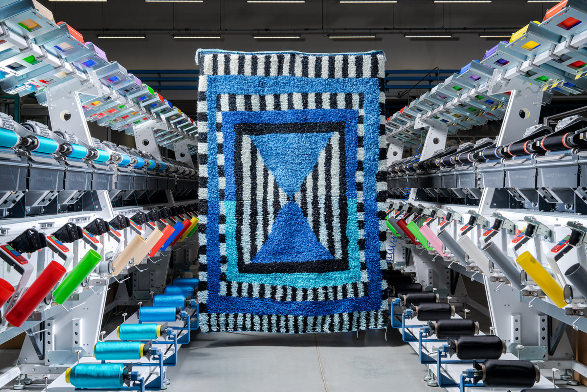 Tappeto in seta blu e bianco con due triangoli al centro, fra le macchine della tessitura Taroni