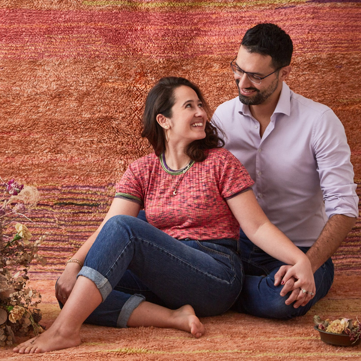 coppia seduta su tappeto dai toni rossi e roda che si tiene la mano e si sorride
