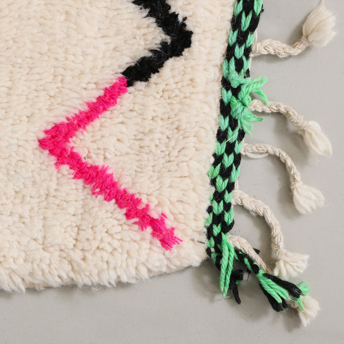 angolo di un tappeto in lana color avorio con  riga spezzata rosa fluo e nera e frangia a scacchi verde e nera