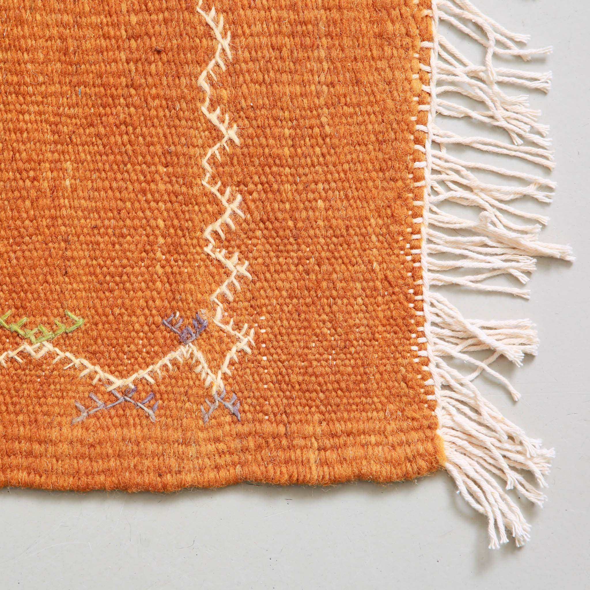Corner detail of handwoven Akhnif Moroccan kilim rug in orange terracotta wool with traditional Amazigh geometric symbols and white fringes, 247x150cm flat-weave carpet from Taznakht region