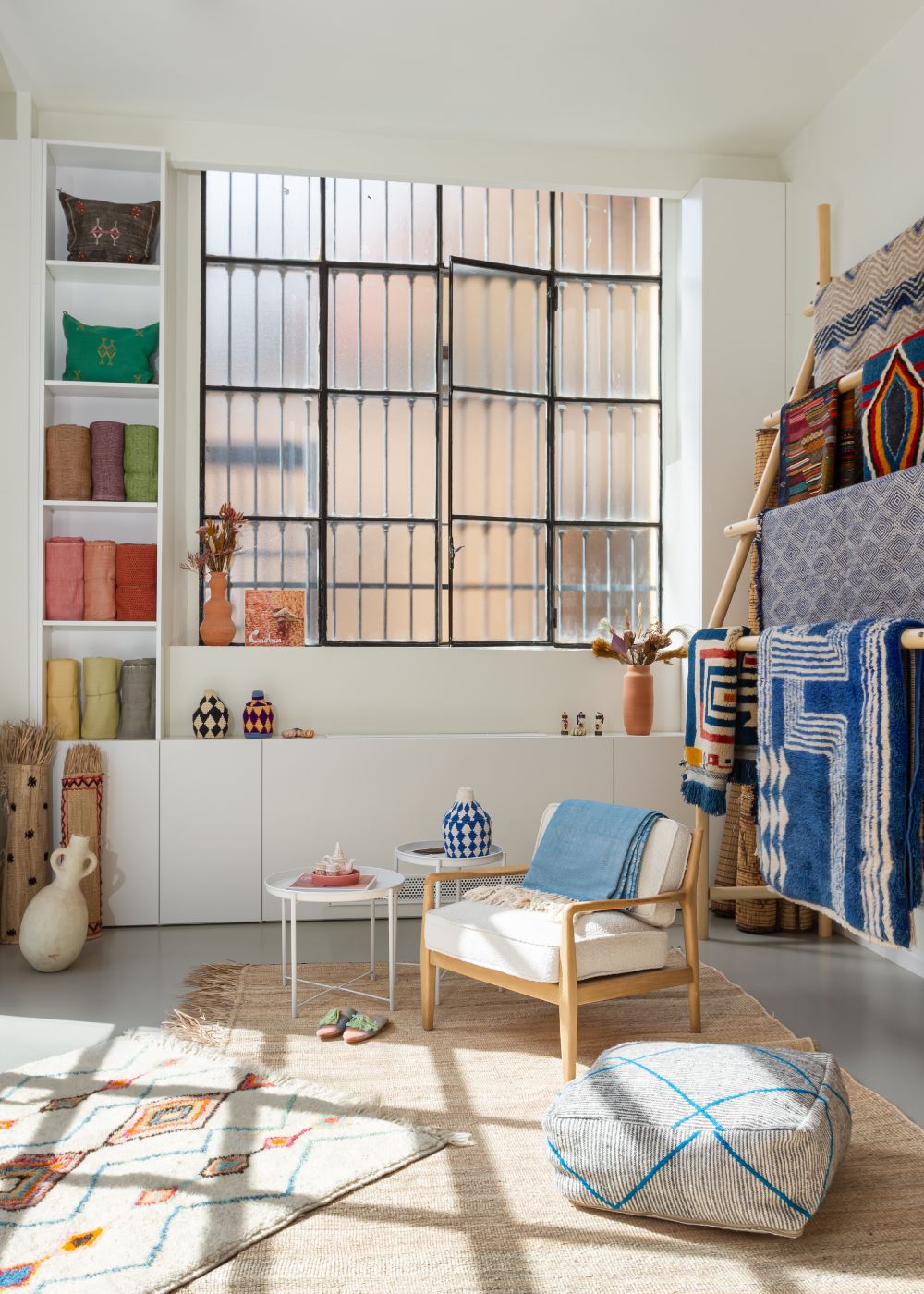 Intérieur de Casa Amar, une salle d'exposition de tapis marocains à Milan. Espace lumineux et aéré présentant des tapis et textiles marocains colorés. De grandes fenêtres éclairent une zone d'exposition élégante avec un fauteuil en bois, des coussins de sol à motifs et un présentoir à tapis. Des étagères garnies de bobines de fil et d'objets décoratifs soulignent l'authenticité de l'artisanat marocain dans cette boutique soigneusement agencée à Milan.