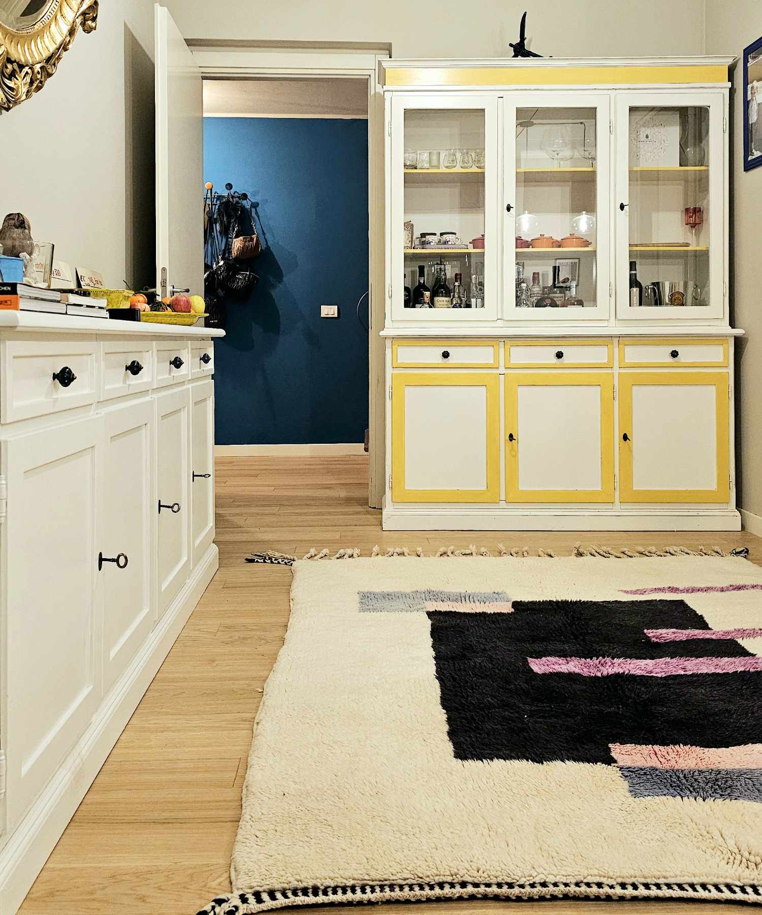 Tapis Beni Mrirt ivoire avec motifs géométriques noirs et roses qui embellit une cuisine de style classique contemporain, vintage, éclectique et boho chic, meublée d'un vaisselier blanc et jaune et d'un mur bleu