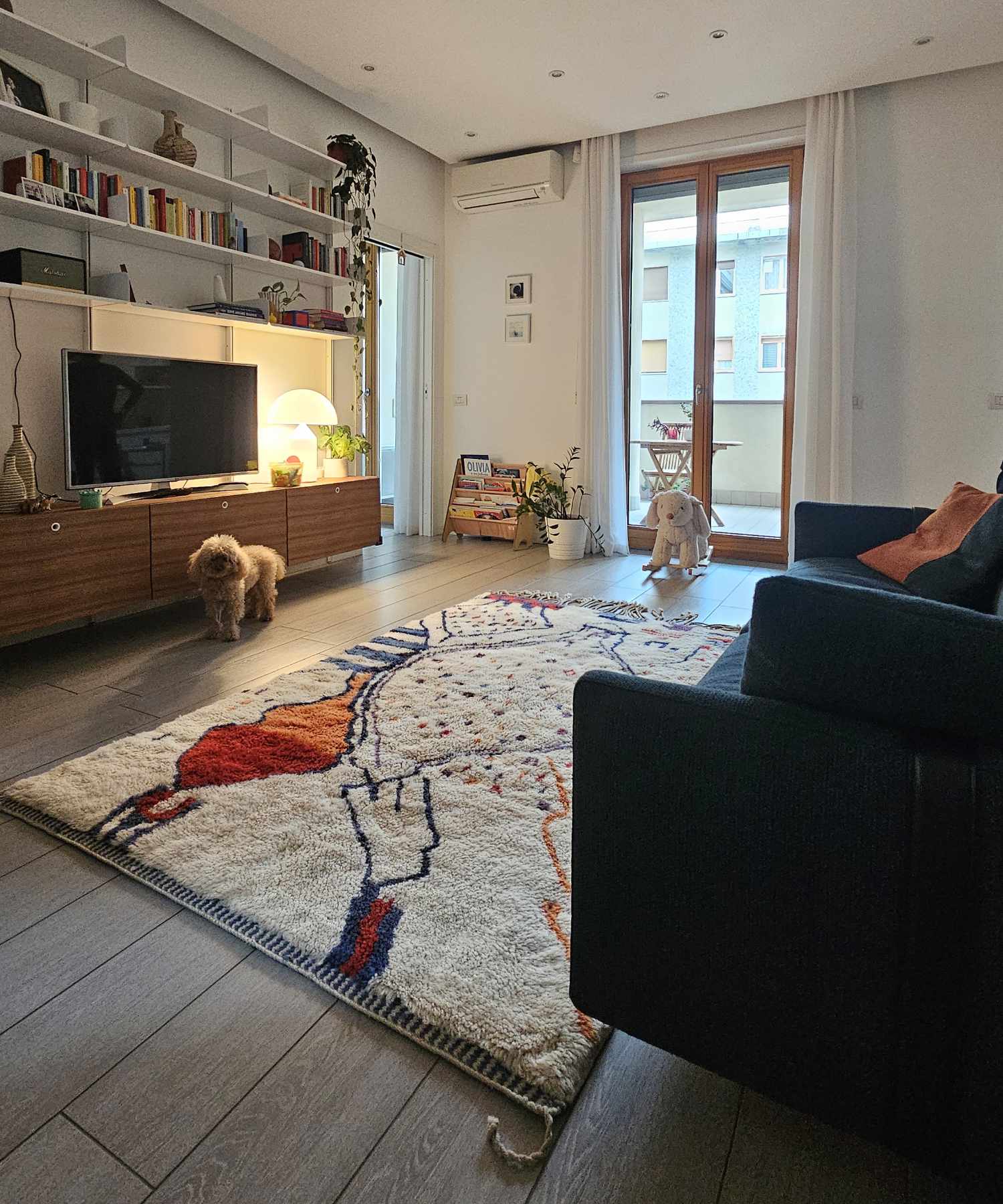 Tapis Beni Mrirt ivoire avec motif abstrait orange et bleu qui décore un salon de style contemporain, scandinave, moderne et minimaliste, avec bibliothèque murale et meuble TV en bois