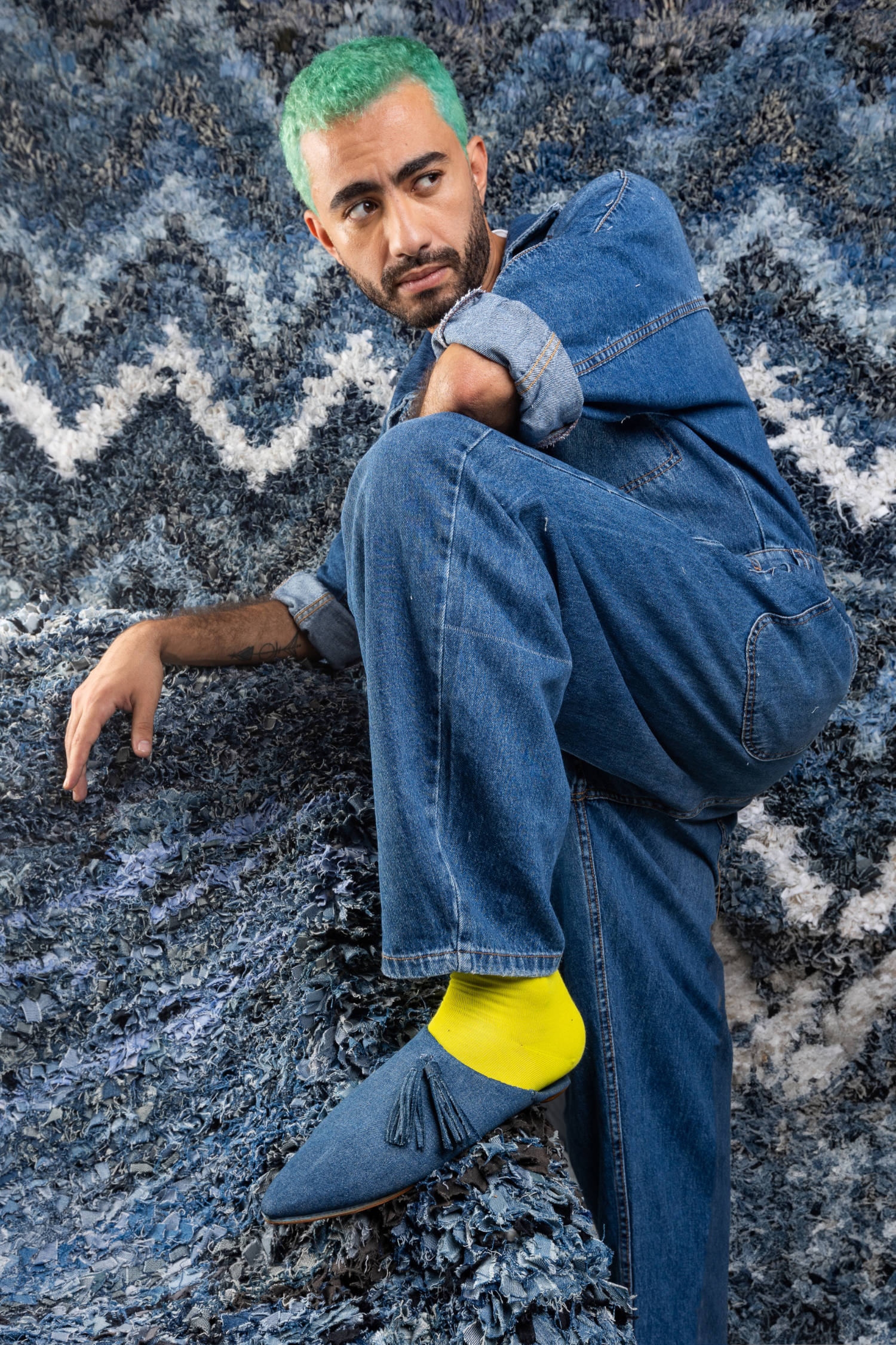 modello in piedi su tappeti boucherouite in denin con una salopette in denim un calzino giallo e una babouche in demin con pompon
