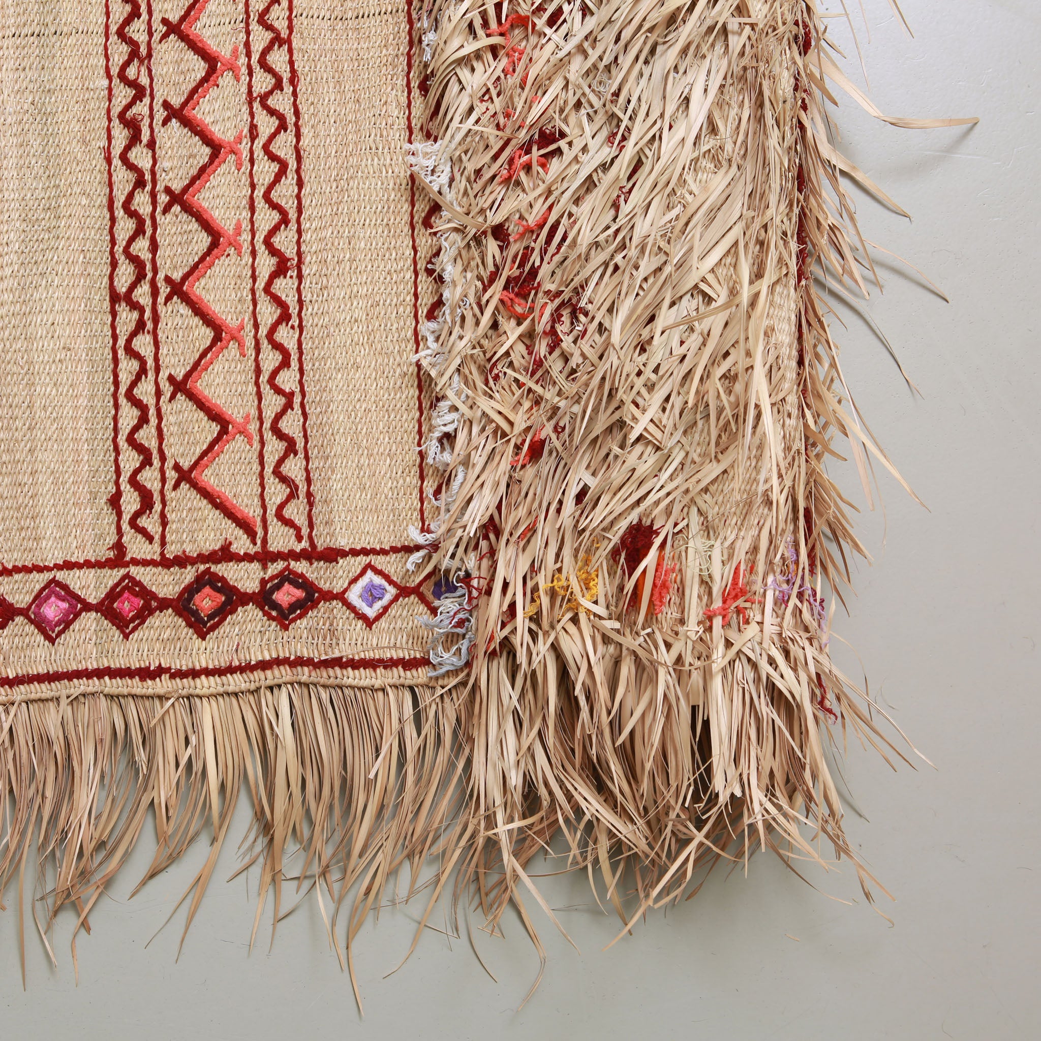 Dettaglio dell'angolo di una stuoia hassira marocchina con ricami geometrici in lana rossa, motivi a rombo colorati e lussureggianti frange in paglia di palma naturale intrecciata a mano