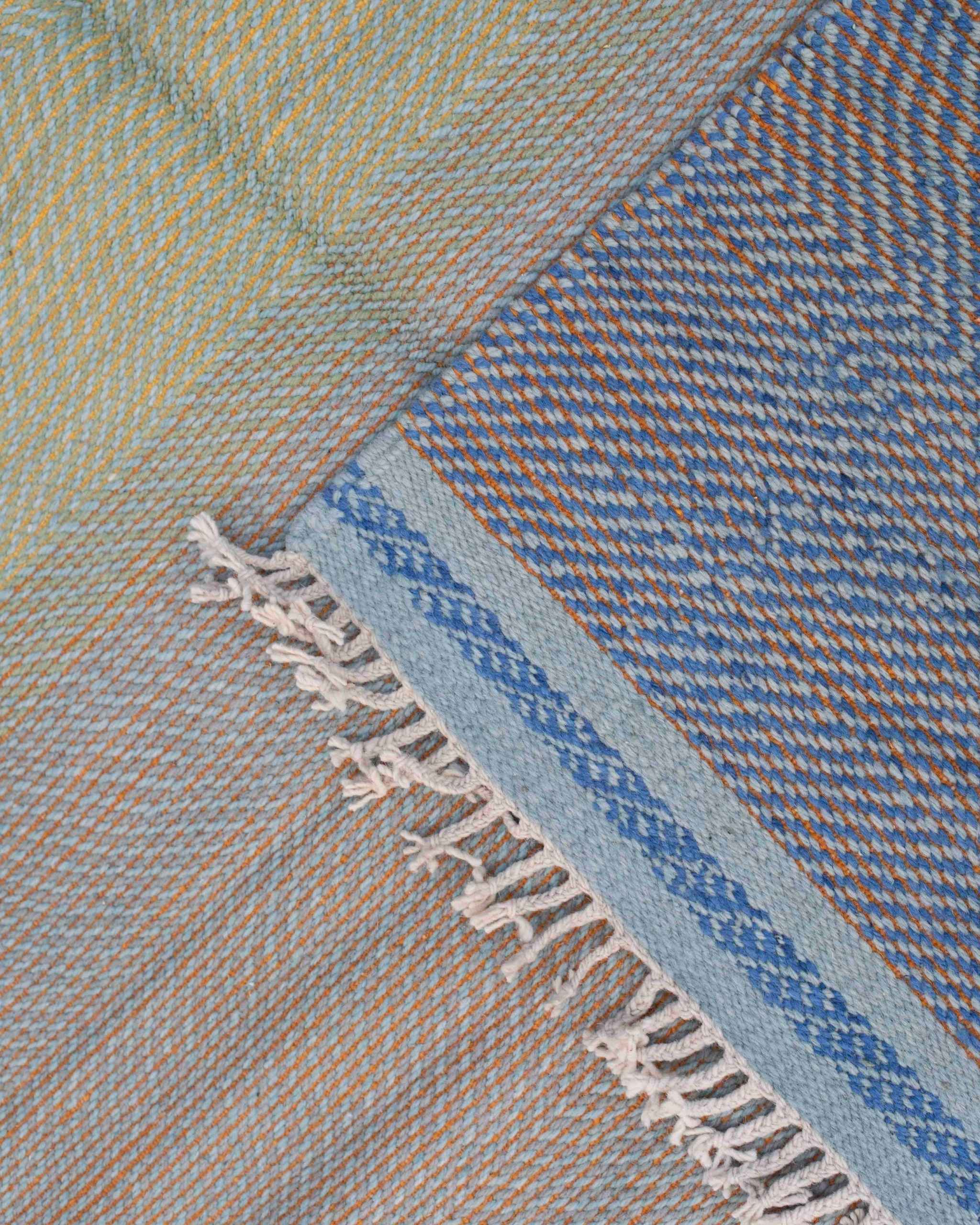 Dettaglio frange e bordura decorativa del tappeto Zanafi in lana, tessitura piatta nei toni azzurro, giallo e arancione