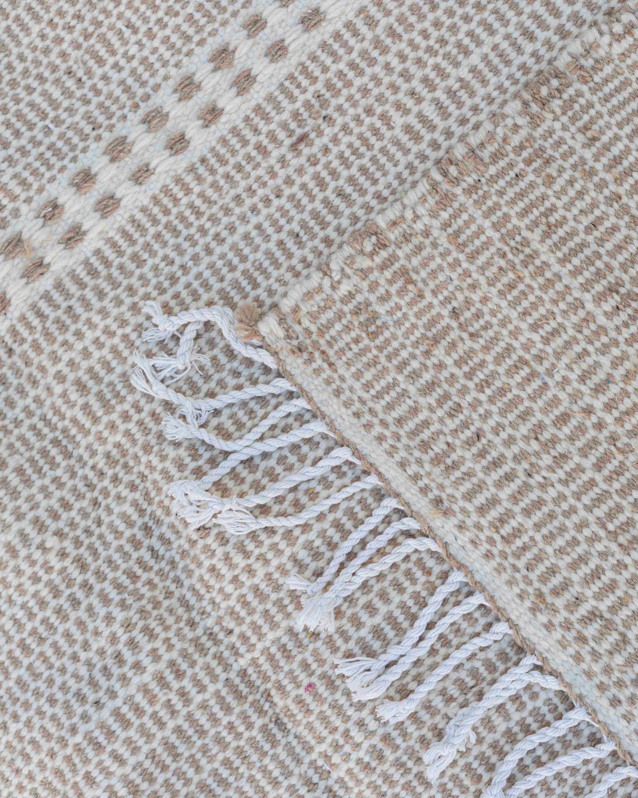 Dettaglio delle frange chiare e della tessitura piatta beige e bianca del tappeto Hanbel