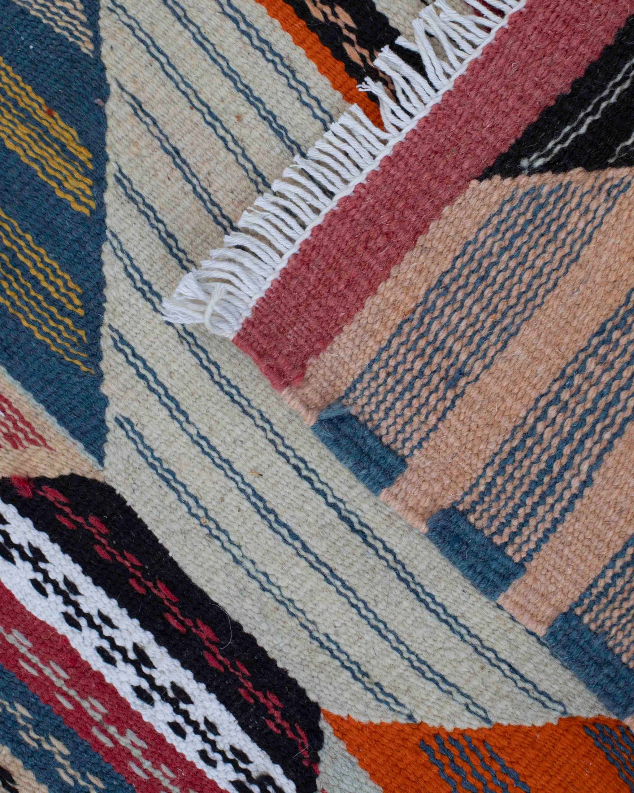Dettaglio del tappetino kilim marocchino in lana con frange e motivo geometrico a righe e losanghe nei toni di rosa, blu, giallo, nero e arancione