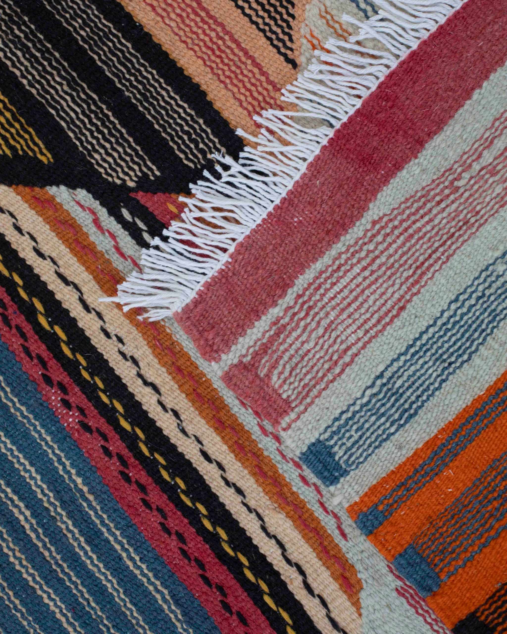 Dettaglio del piccolo tappeto kilim Kharita in lana a losanghe e righe arancione, rosa, blu, crema, nere con frange bianche