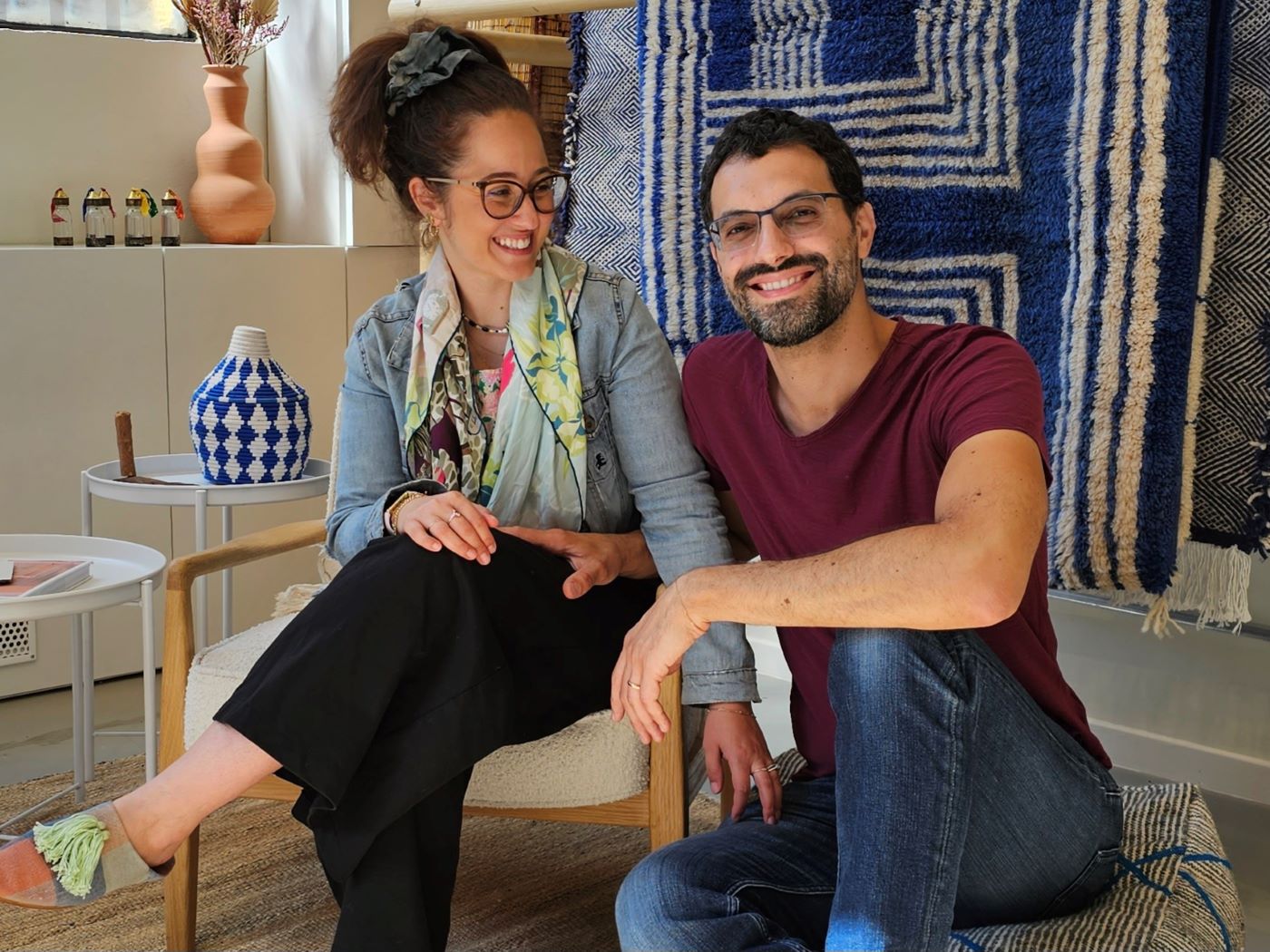 Les fondateurs de Casa Amar, Jihane et Matteo, souriants dans leur showroom de tapis marocains. Le couple est assis sur des chaises design, entouré de vibrants tapis marocains bleus, de poteries artisanales et de décoration bohème, illustrant leur mélange d'artisanat traditionnel marocain et de design d'intérieur moderne