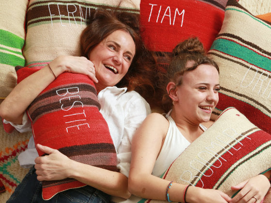 Collection Entre les Lignes: Sabina Maffei Plazner et sa fille Costanza Marin sourient allongées sur une composition de coussins artisanaux fabriqués à partir de couvertures vintage marocaines en laine et de tissus raffinés italiens. Les coussins, servant à la fois de fond et de sujet, sont brodés à la main avec des textes personnalisés.