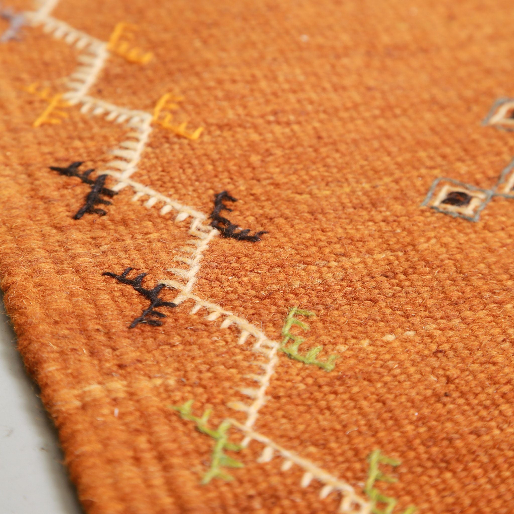 Close-up of traditional Amazigh geometric symbols on orange Akhnif Moroccan rug showing black, blue and green embroidered motifs on flat-weave kilim texture