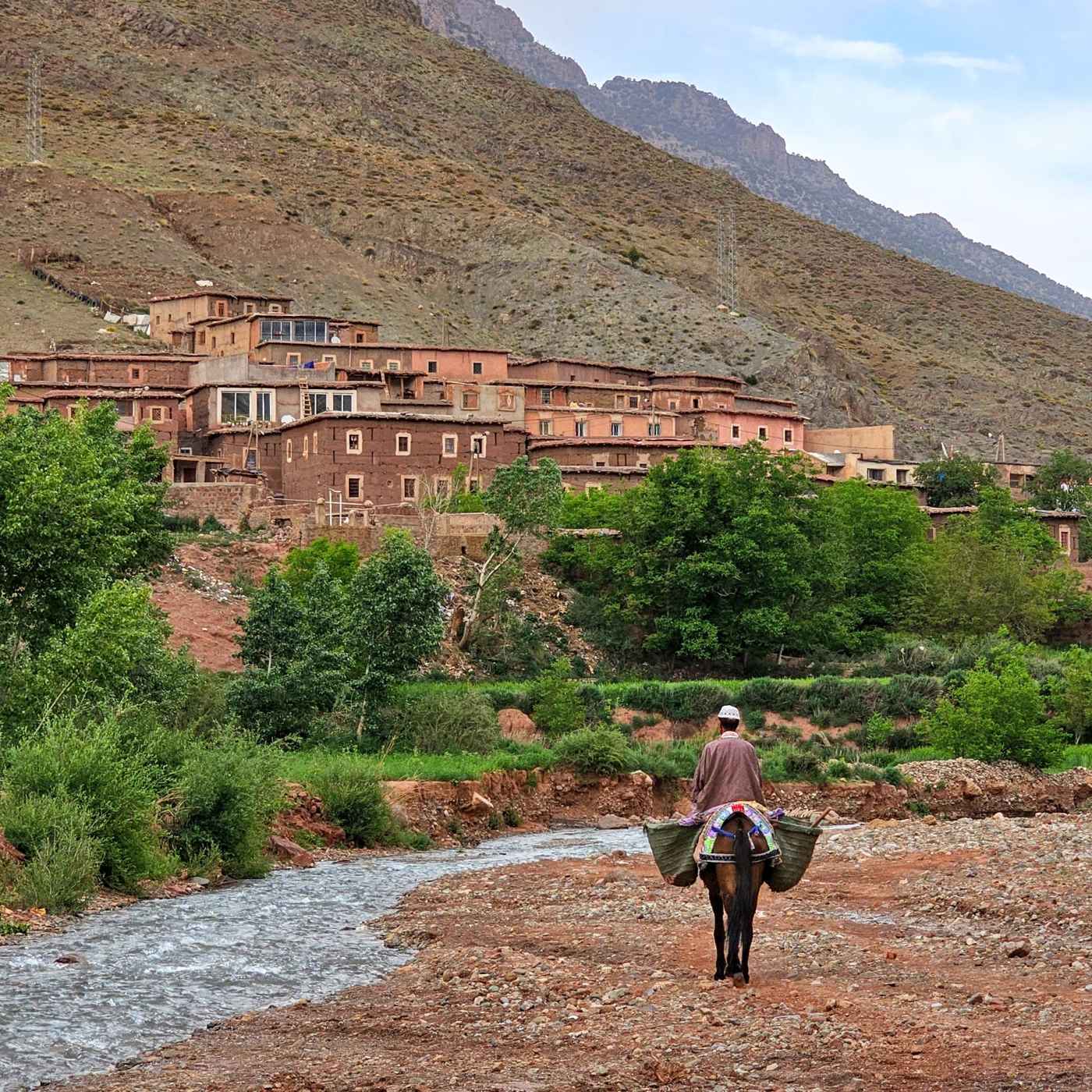 Sustainable traditional village in Atlas Mountains, Morocco: mud-brick houses, eco-friendly local transport along river, Berber architecture integrated into mountain landscape