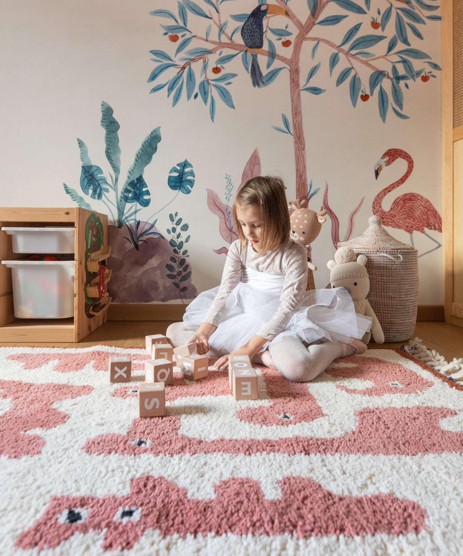 Bambina gioca con cubi educativi sul tappeto Animauì rosa, lana marocchina annadata a mano, ambientazione cameretta con murales naturalistici