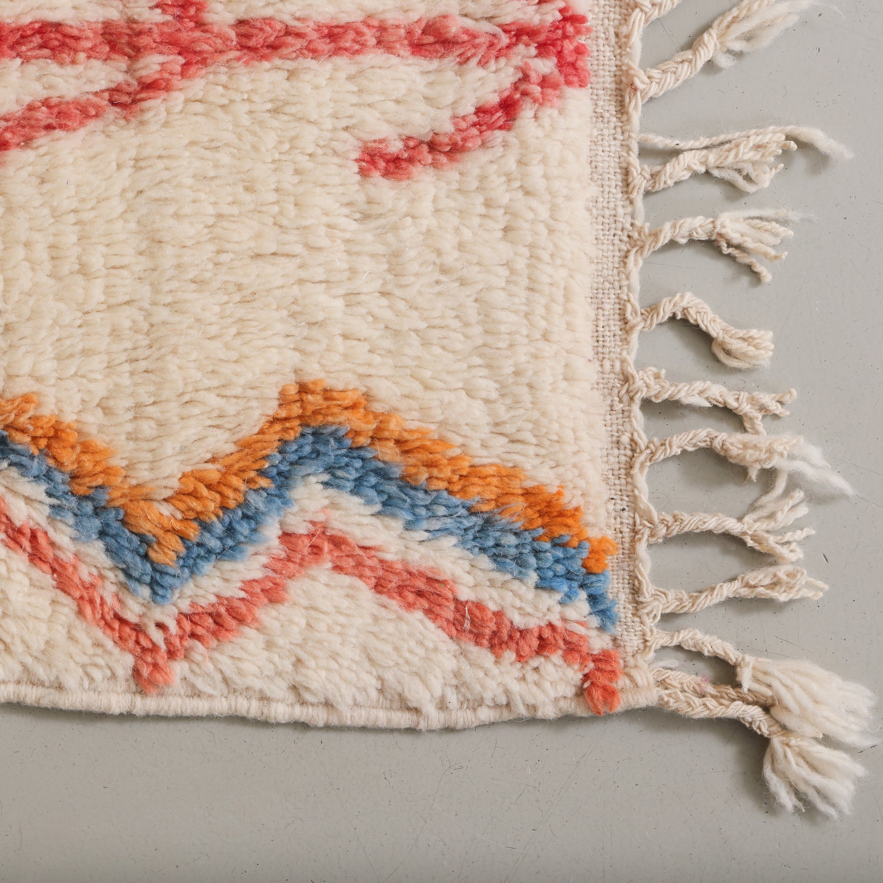 dettaglio dell'angolo di un tappeto. si possono vedere delle linee spezzate affiancate di diversi colori e la frangia con corte trecce bianche