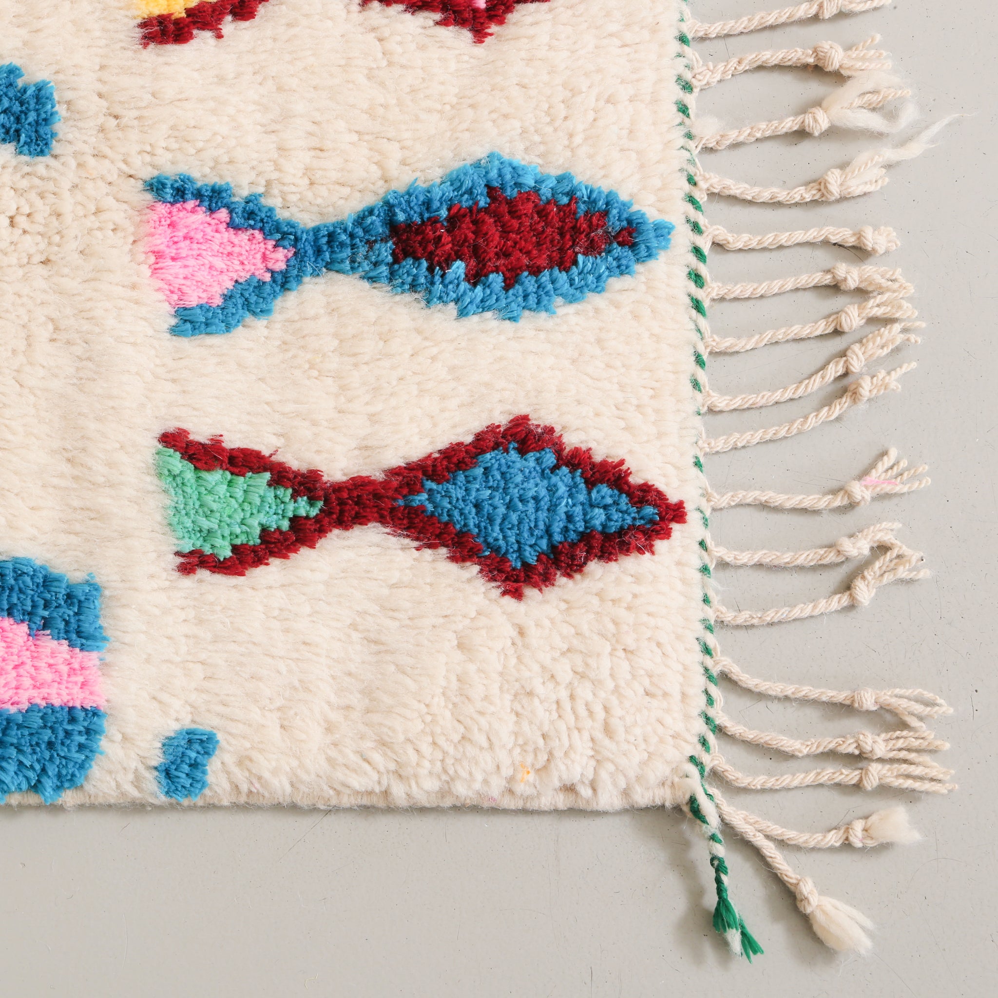 dettaglio dell'angolo di un tappeto in lana annodata con simboli che ricordano dei pesci di colore azzurro bordeaux e rosa. il tappetto è stato chiuso con una treccia bianca e verde. la frangia è sottile e lunga