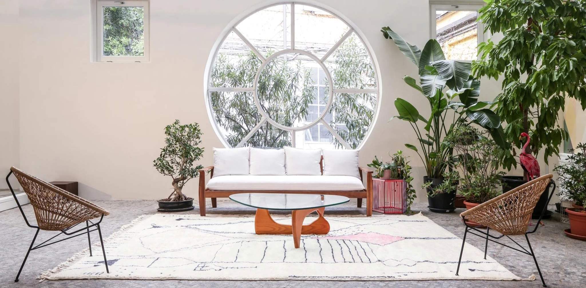 Tappeto berbero Beni Mrirt autentico in lana naturale che arreda uno studio di architettura contemporaneo. Design minimalista con divano bianco, sedie in rattan e tavolino Noguchi, illuminato da finestra circolare e vegetazione mediterranea.