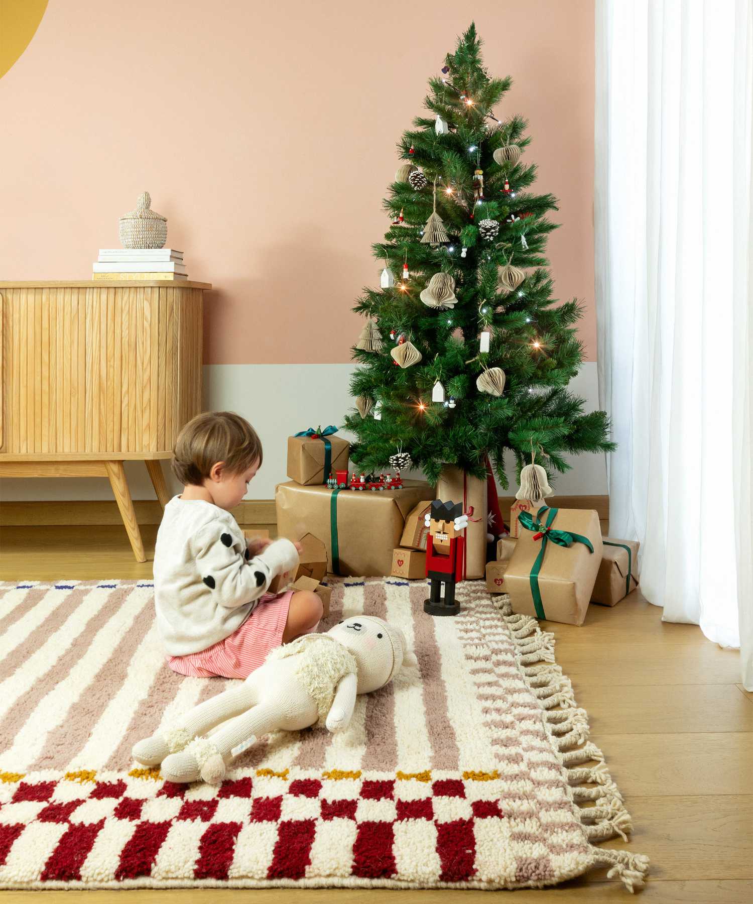 Bambina seduto sul tappeto Linhas in lana marocchina con motivi rosa e rossi, momento natalizio con albero decorato e regali, atmosfera festiva e accogliente