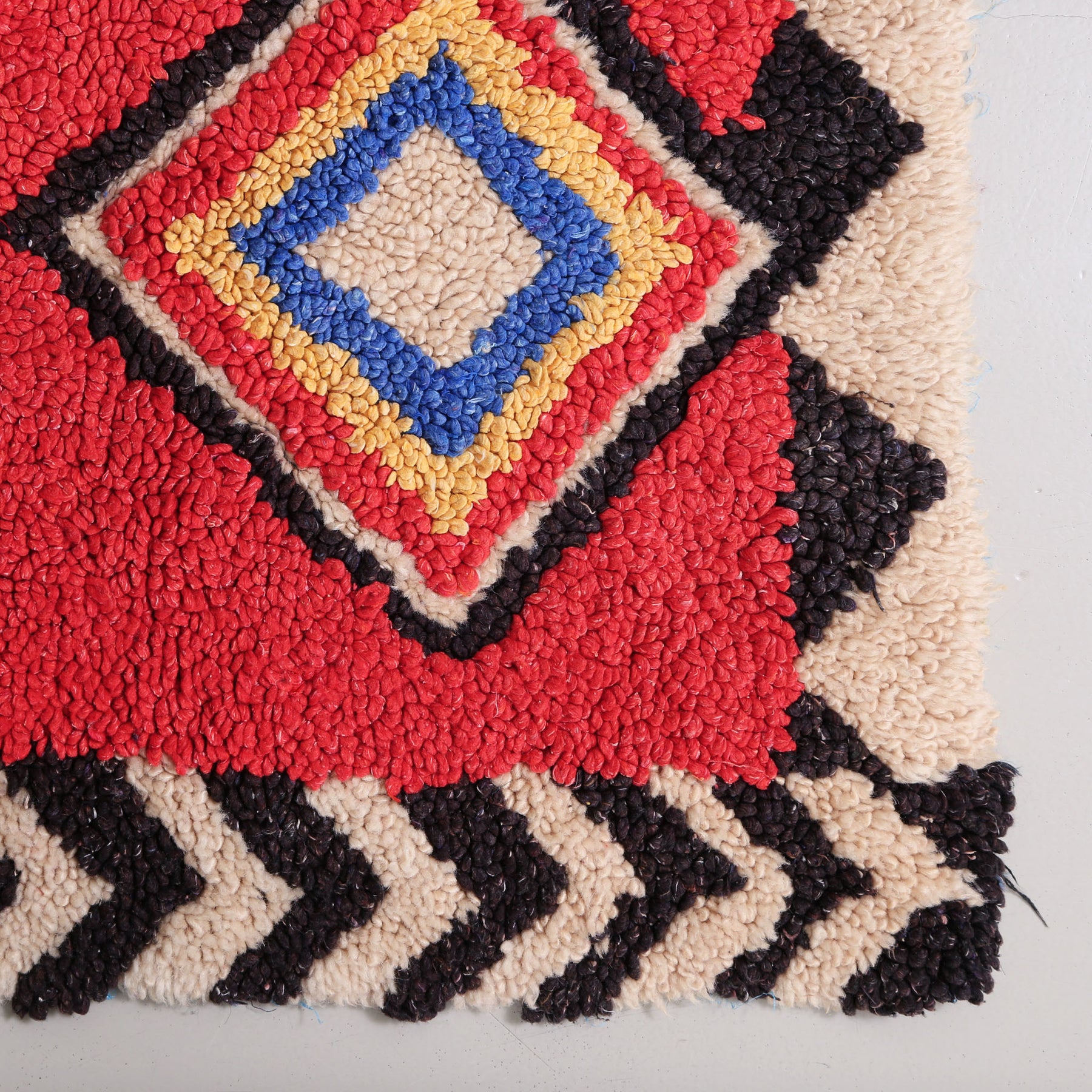 angolo di un tappeto zindeck realizzato annodando lana colorato si un sacchetto di plastica. base lana rossa con rombo colore nero bianco rosso giallo blu e bianco. i bordi sono bianchi e neri