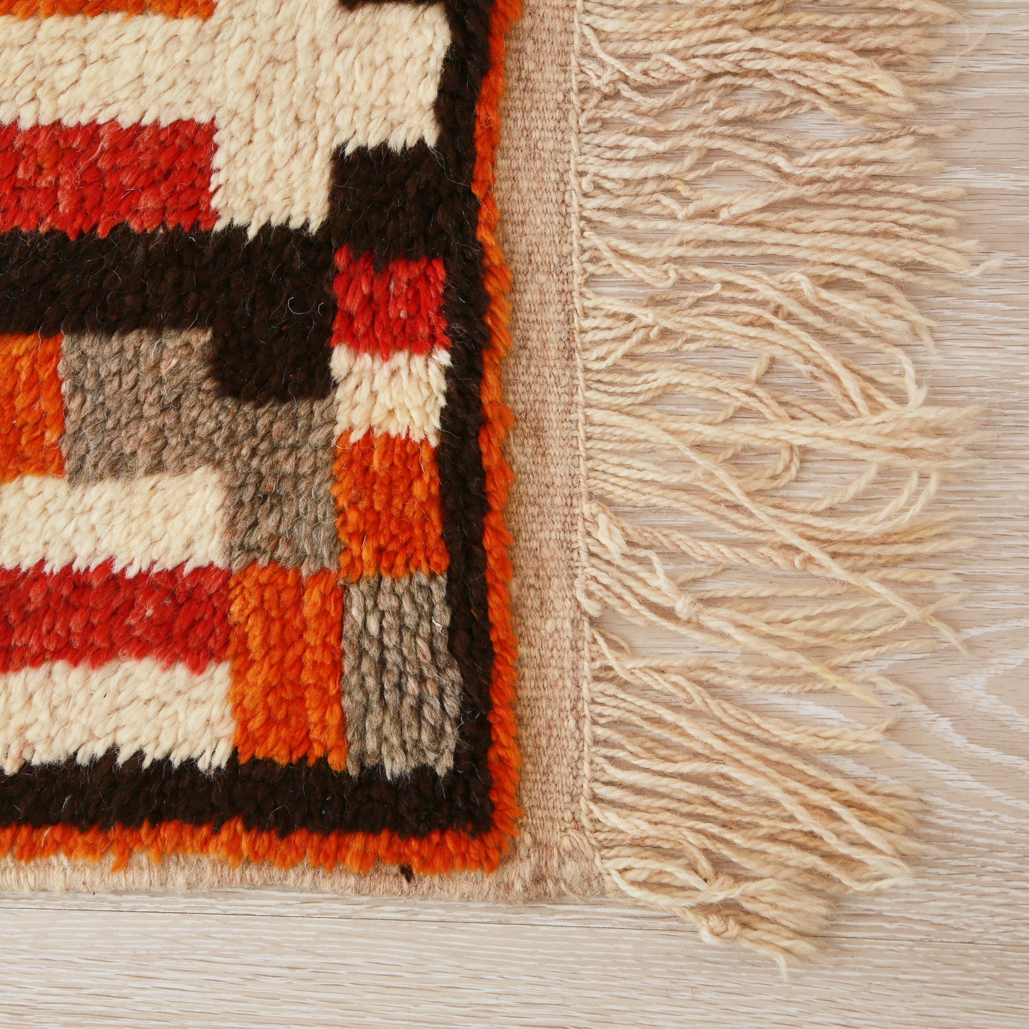 angolo di un tappeto stile azilal con quadrati e rettangoli di diversi colori, come diverse sfuture di rosso, arancione e grigio, con un lunga frangia sottile bianca