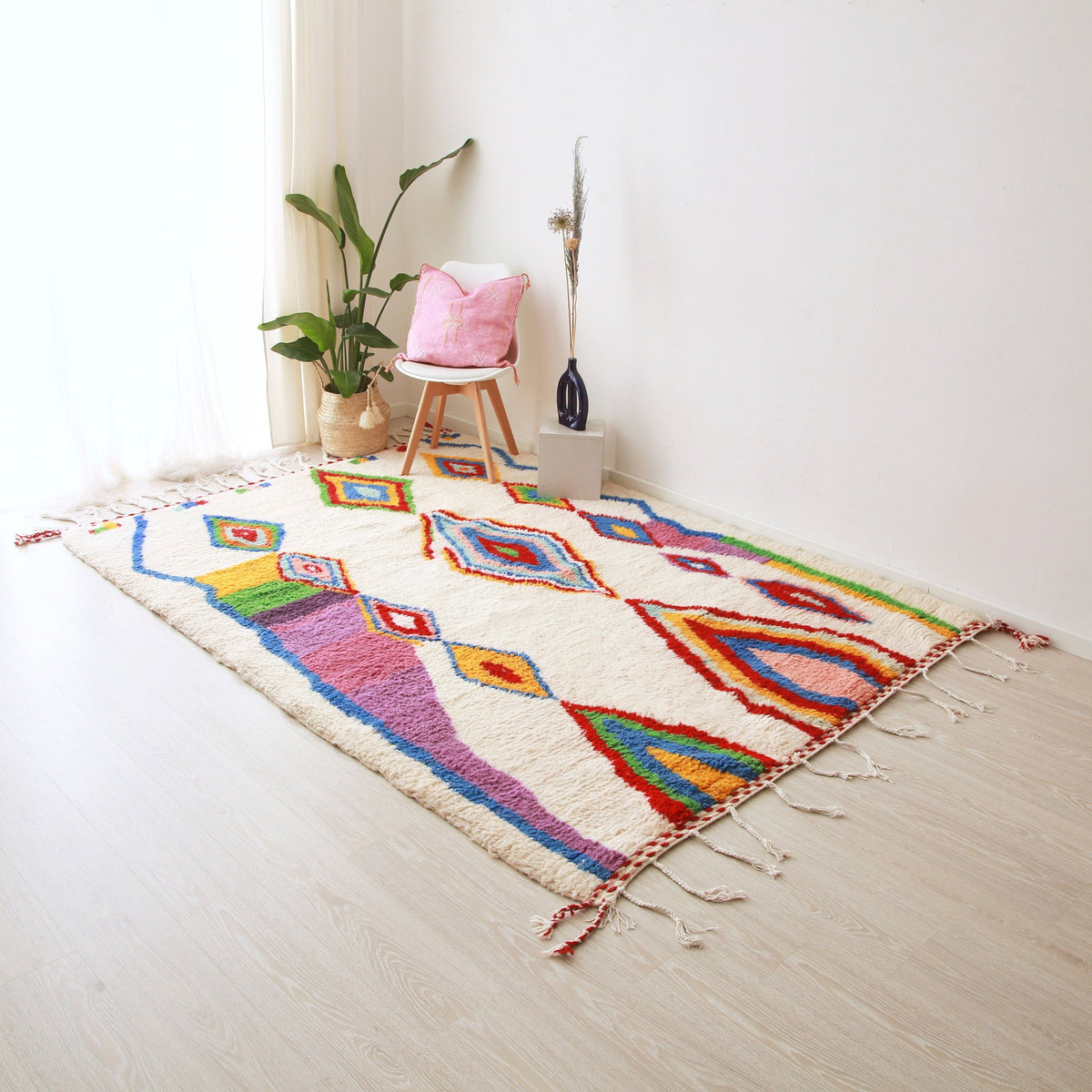 tappeto azilal disteso realizzato con lana bianca e rombi di diverse dimensioni dai colori brillanti. Sul tappeto c'è una pianta, una sedia bianca con un cuscino in sabra rosa e un vaso in ceramica blu