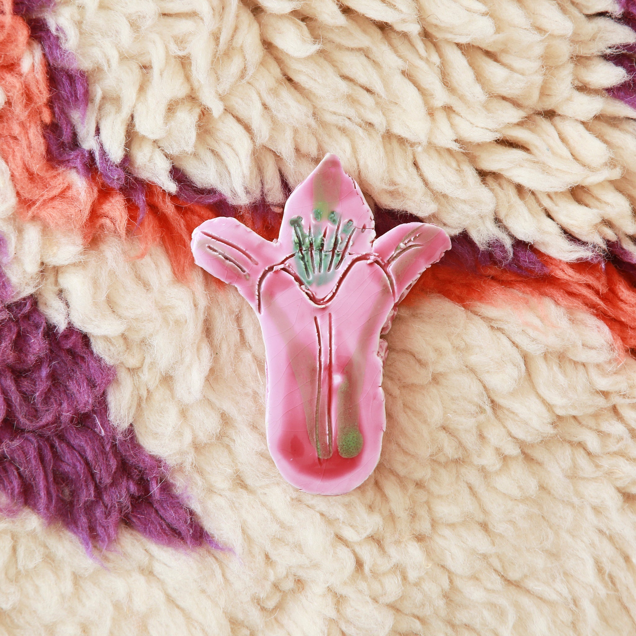 fiore rosa realizzato in ceramica appossiato su un tappeto di lana bianca e con accenni di viola e arancione