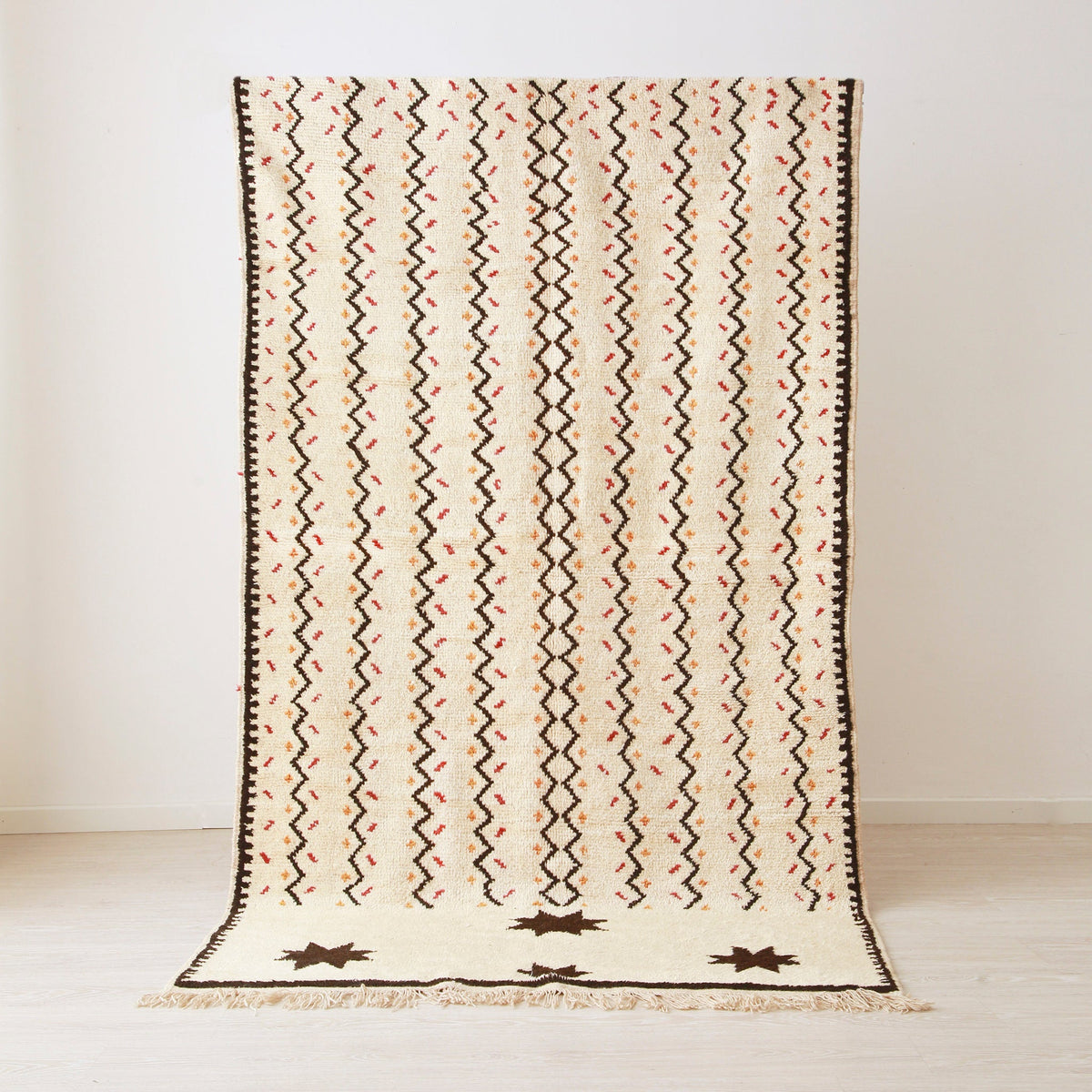 tappeto stile azilal appeso realizzato su base bianche con linee a zig zag nere e quattro stelle nere. Ai lati delle linee spezzate delle piccole linee arancioni e rosse