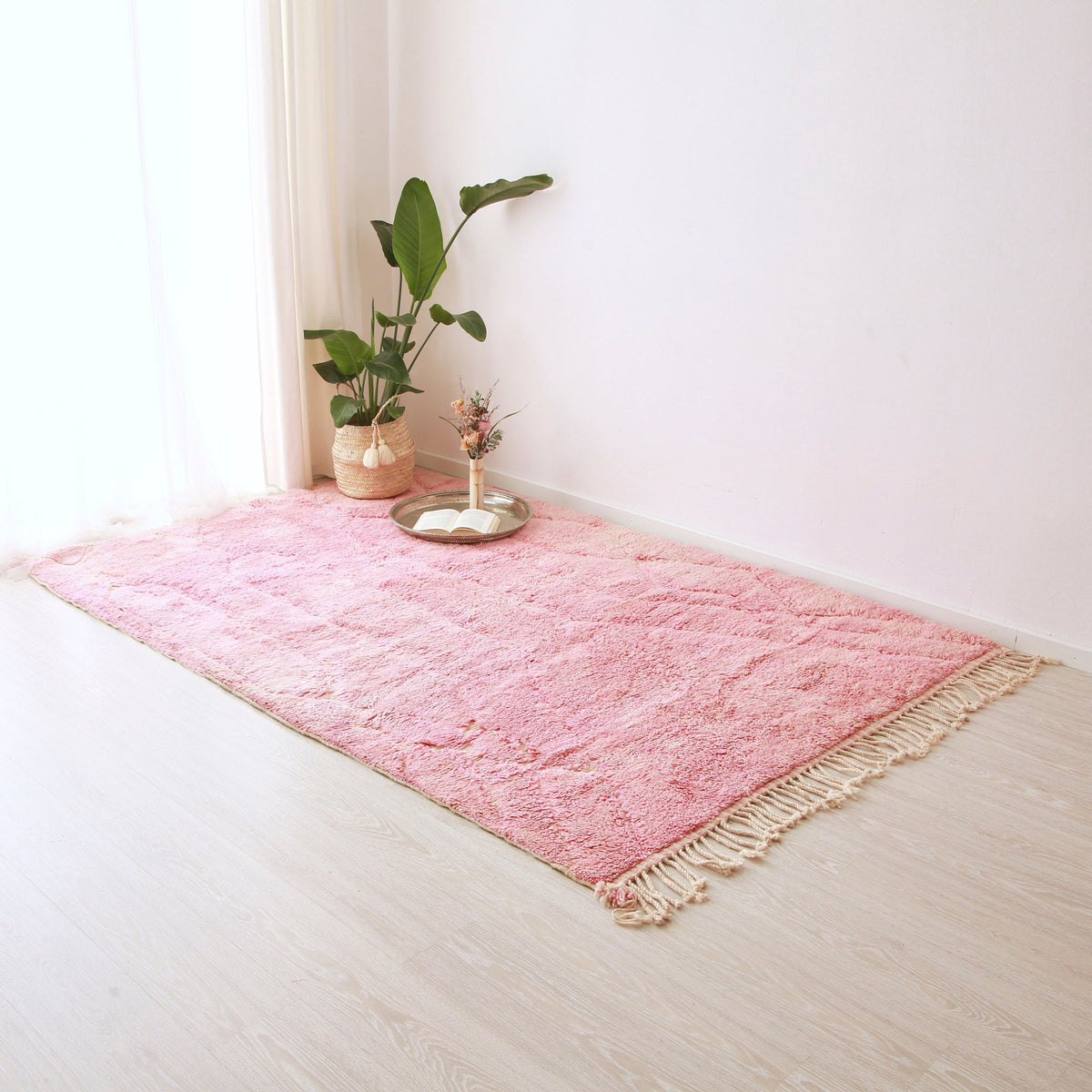 Tappeto beni mrirt di colore rosa e con un lunga frangia disteso sul pavimento. Sul tappeto sono presenti un pianta e un patto in argento con sopra un libro aperto e una composizione floreale