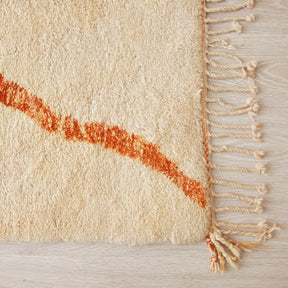 primo piano dell'angolo di un tappeto beni mrirt per mettere in risalto la frangia. Un linea di lana arancione taglia diagonlmente l'immagine per terminare sull'angolo del tappeto