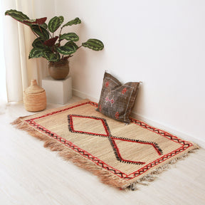 tappeto stuoia hassira in paglia piccola con ricami geometrici in lana neri rossi a forma di rombi e cornice laterale disteso sul pavimento con un cuscino in sabra color liquirizia sopra